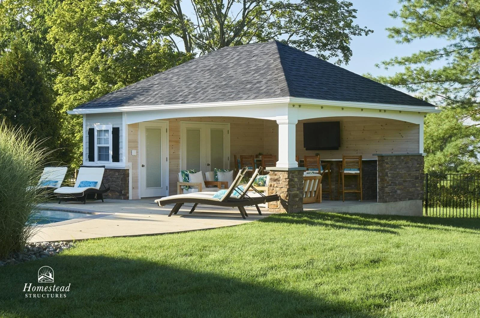 A backyard with a poolside pavilion featuring an outdoor living space, bar area, and a TV mounted on the wooden wall, surrounded by a well-manicured lawn and tall trees.