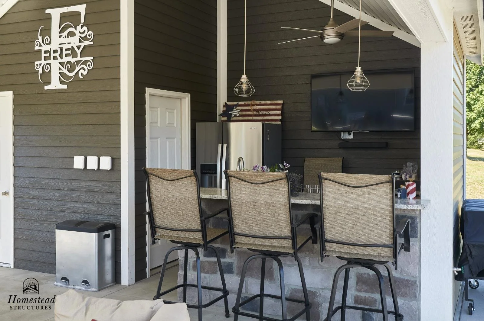 Backyard outdoor kitchen with a bar with four chairs, a refrigerator, a large television mounted on the wall, and decorative American flag and star decorations.