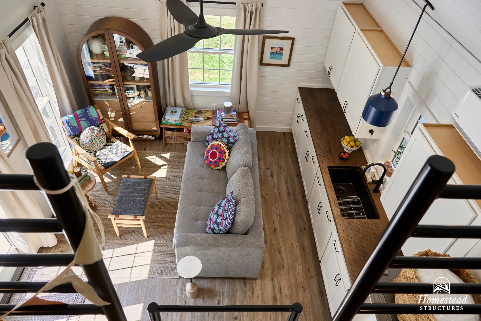 A cozy living room with white wood-paneled walls, a gray sofa with colorful pillows, a wooden armchair with a patterned pillow, a small side table, a beige rug, and a large window with beige curtains. The room has a wooden shelf, a framed picture, an