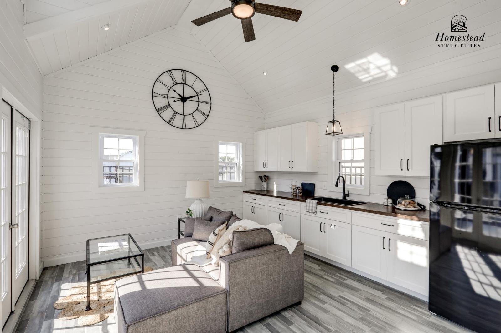 Interior view of a modern, minimalist kitchen and living area with white shiplap walls, small windows, a black wall clock, a ceiling fan, and a gray sectional sofa with pillows, a glass coffee table, and a black refrigerator.