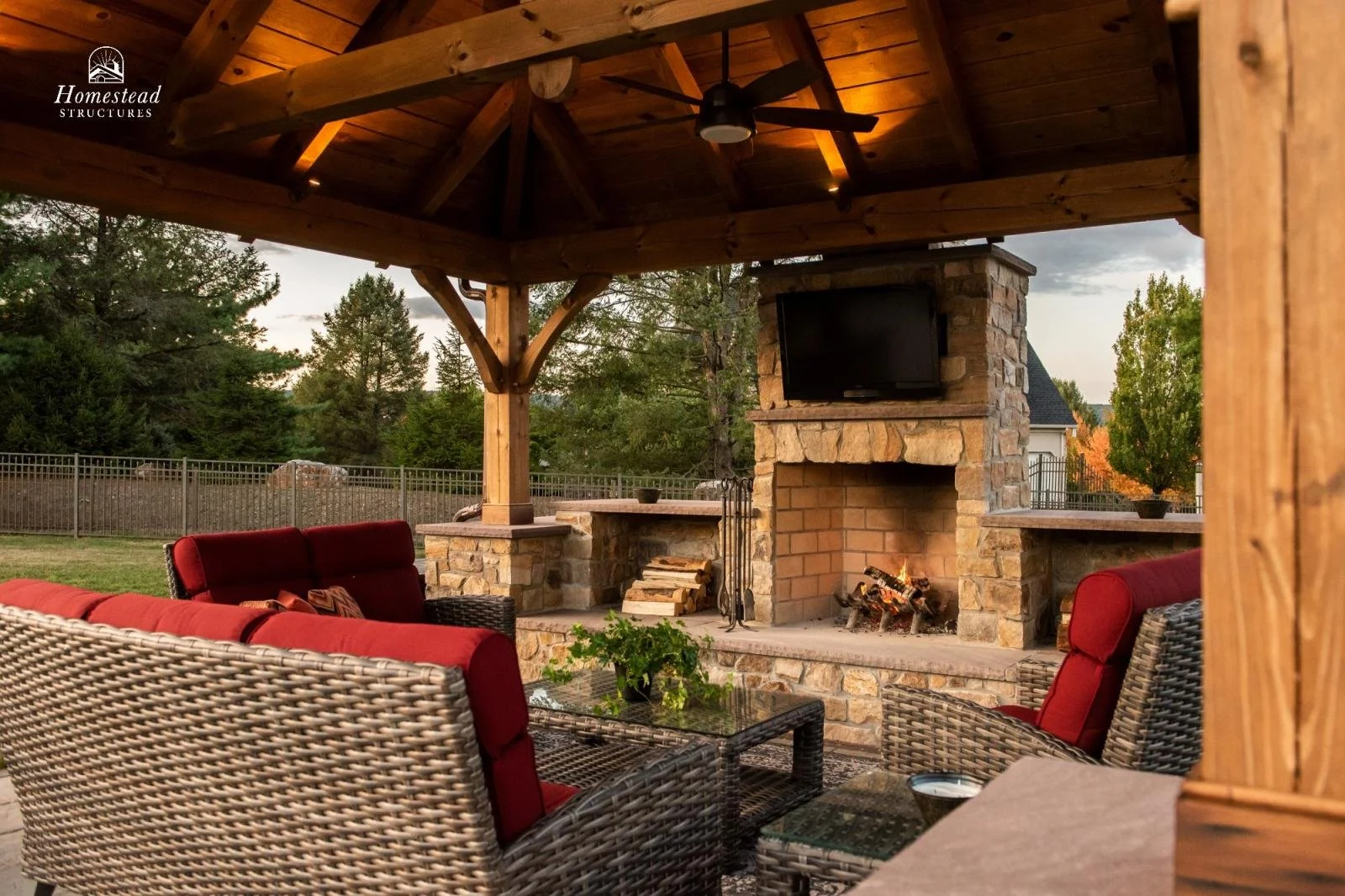 Outdoor covered patio with a stone fireplace, a ceiling fan, and wicker furniture with red cushions. There is a flat-screen TV mounted above the fireplace and trees in the background.