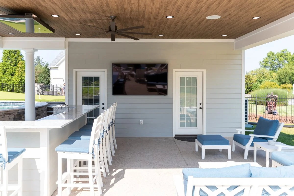 Outdoor patio area with bar counter, bar stools, lounge chairs, a mounted flat screen TV, and doors leading to the backyard with pool and green lawn.