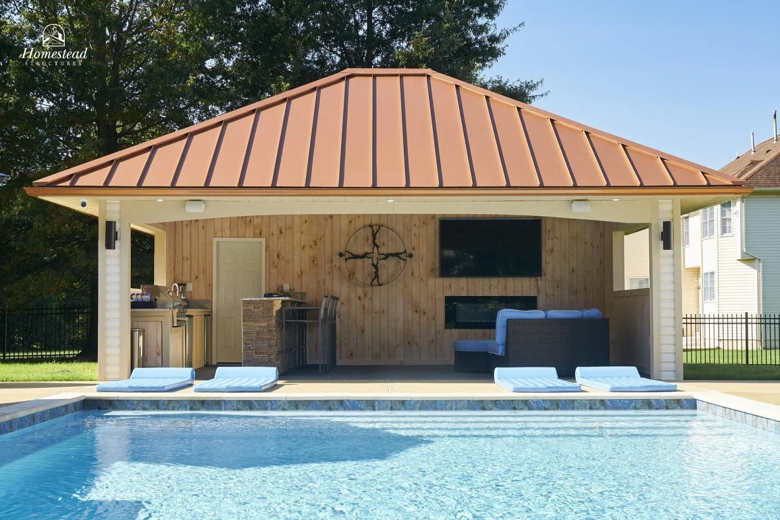 Outdoor pool area with lounge chairs, an open pavilion with a copper roof, a TV, a seating area, a barbecue grill, and a wooden wall with decor, surrounded by a fence and trees.