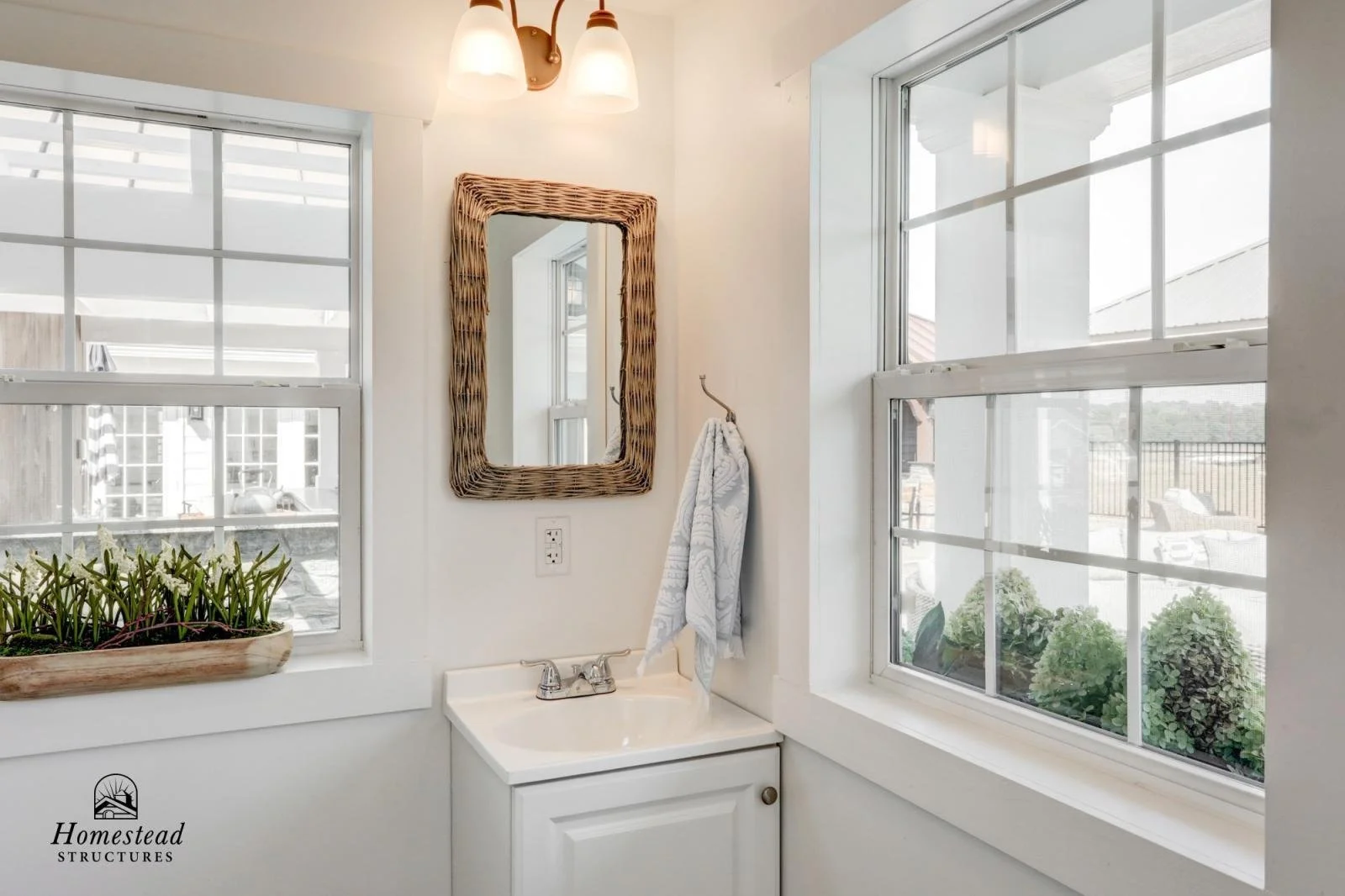 A small bathroom corner with white walls, two large windows with white frames, a small white sink with a silver faucet, a wall-mounted mirror with a woven rattan frame, a light fixture with three white lights, a towel hanging on a hook, a window box 