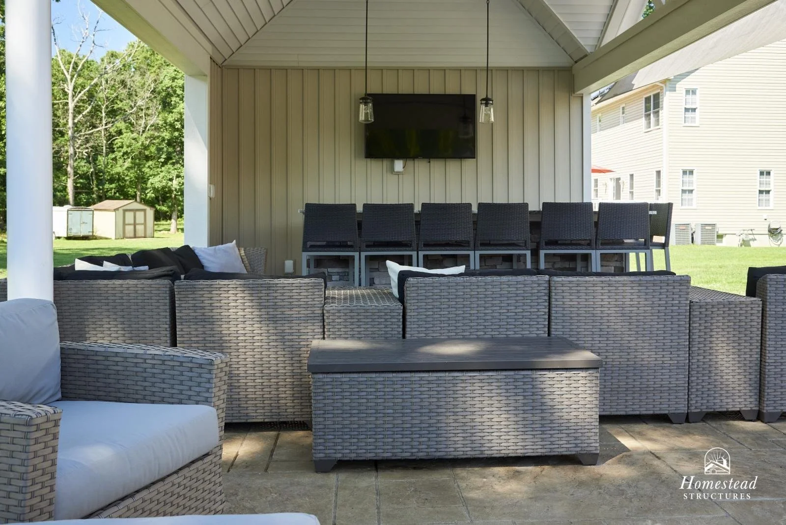 Outdoor patio with woven furniture, cushioned seats, a wall-mounted TV, and bar stools under a covered roof.