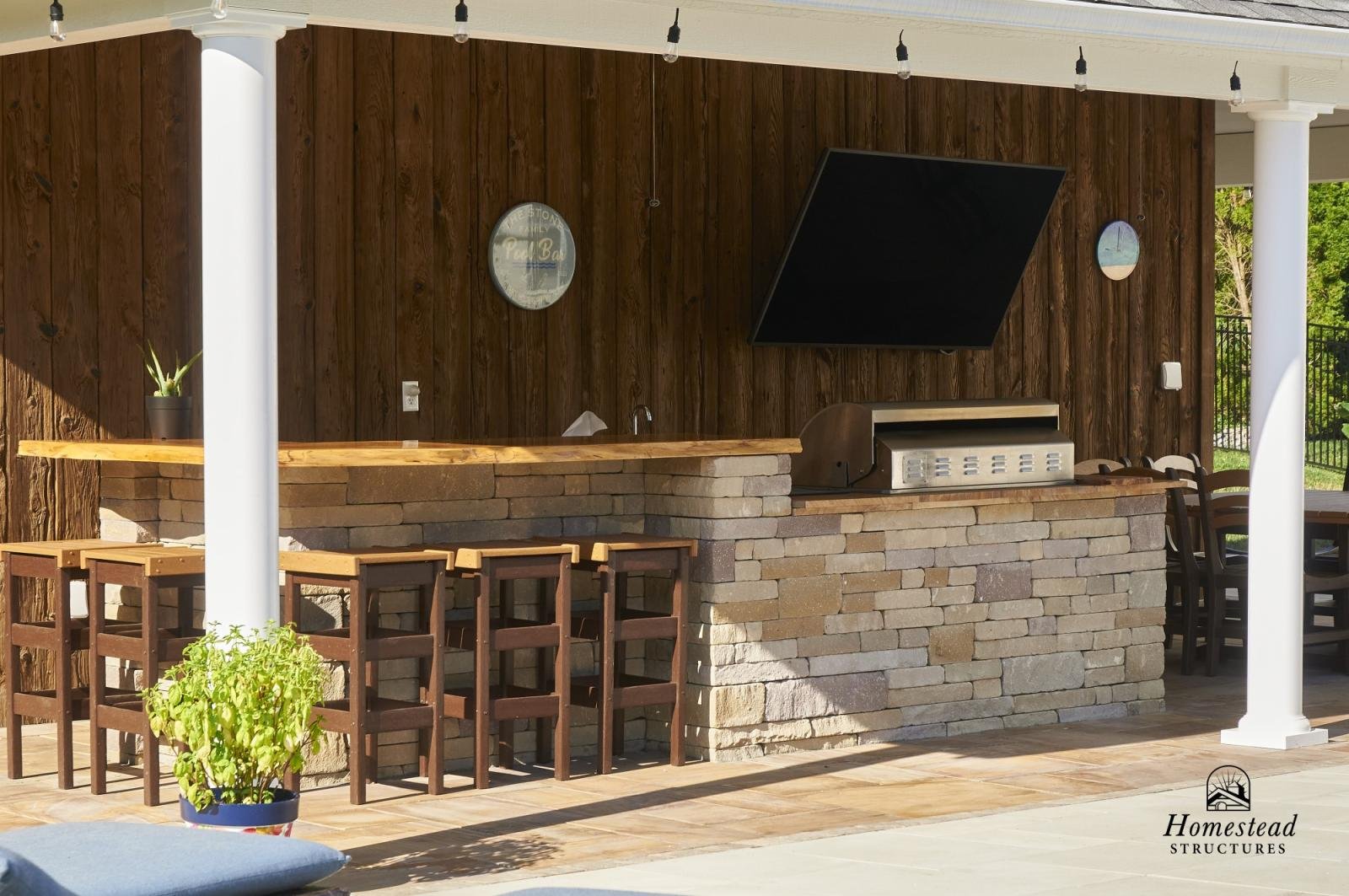 Outdoor bar area with wooden and stone construction, bar stools, a grill, a small sink, and decorative wall mirrors, set on a patio with string lights and greenery.