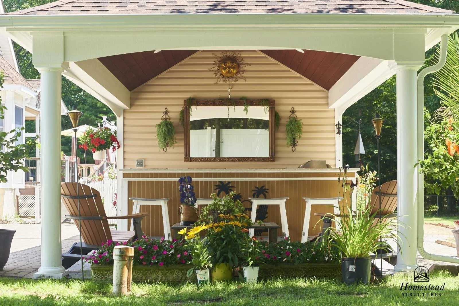 A covered outdoor patio with lounge chairs, potted plants, and a bar area with a mirror, hanging greenery, and decorative lighting. Behind is a green yard and neighboring houses.