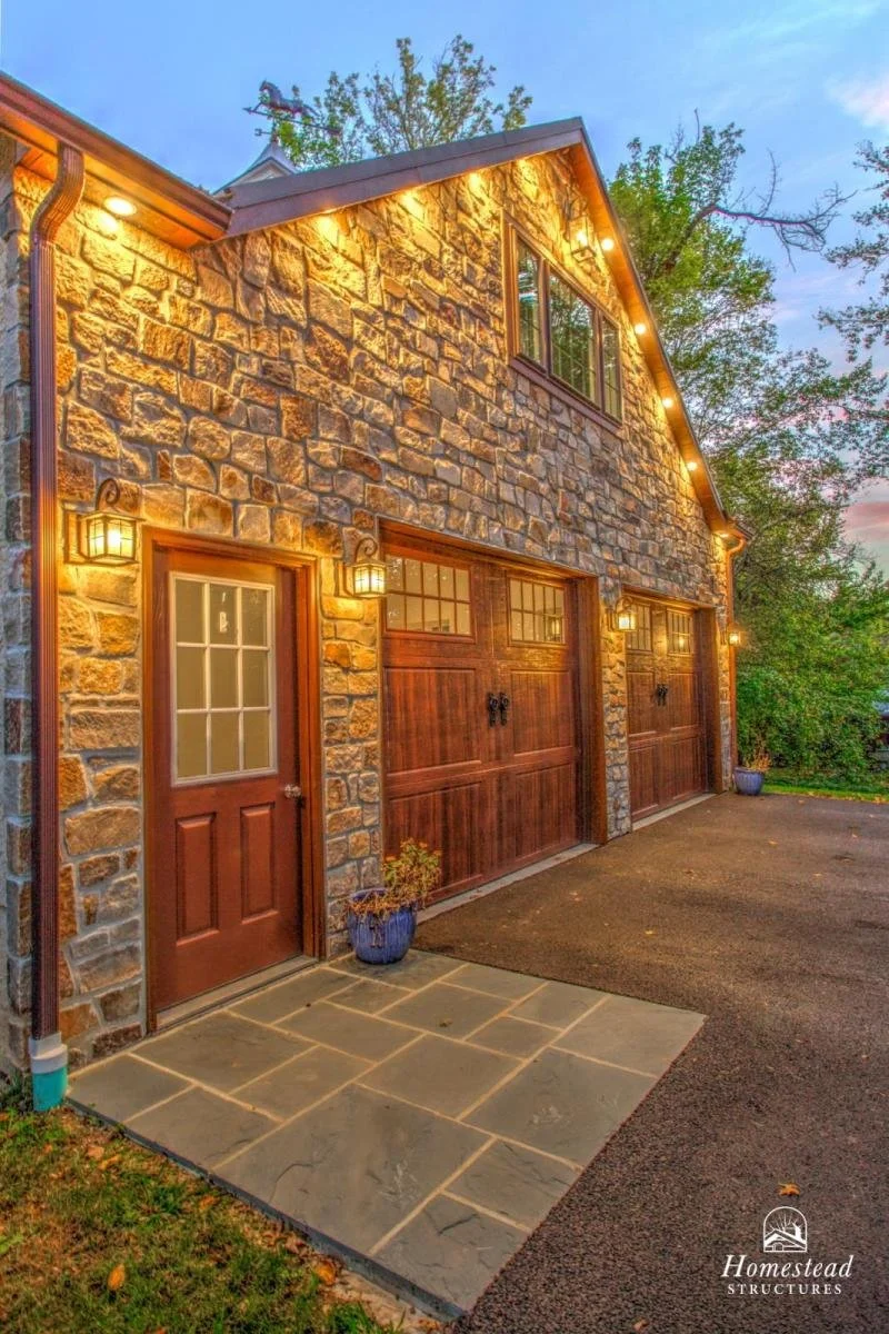 Exterior of a stone and wood barn-style garage with three doors, outdoor lighting, and a small entry door, surrounded by trees with string lights along the roofline.