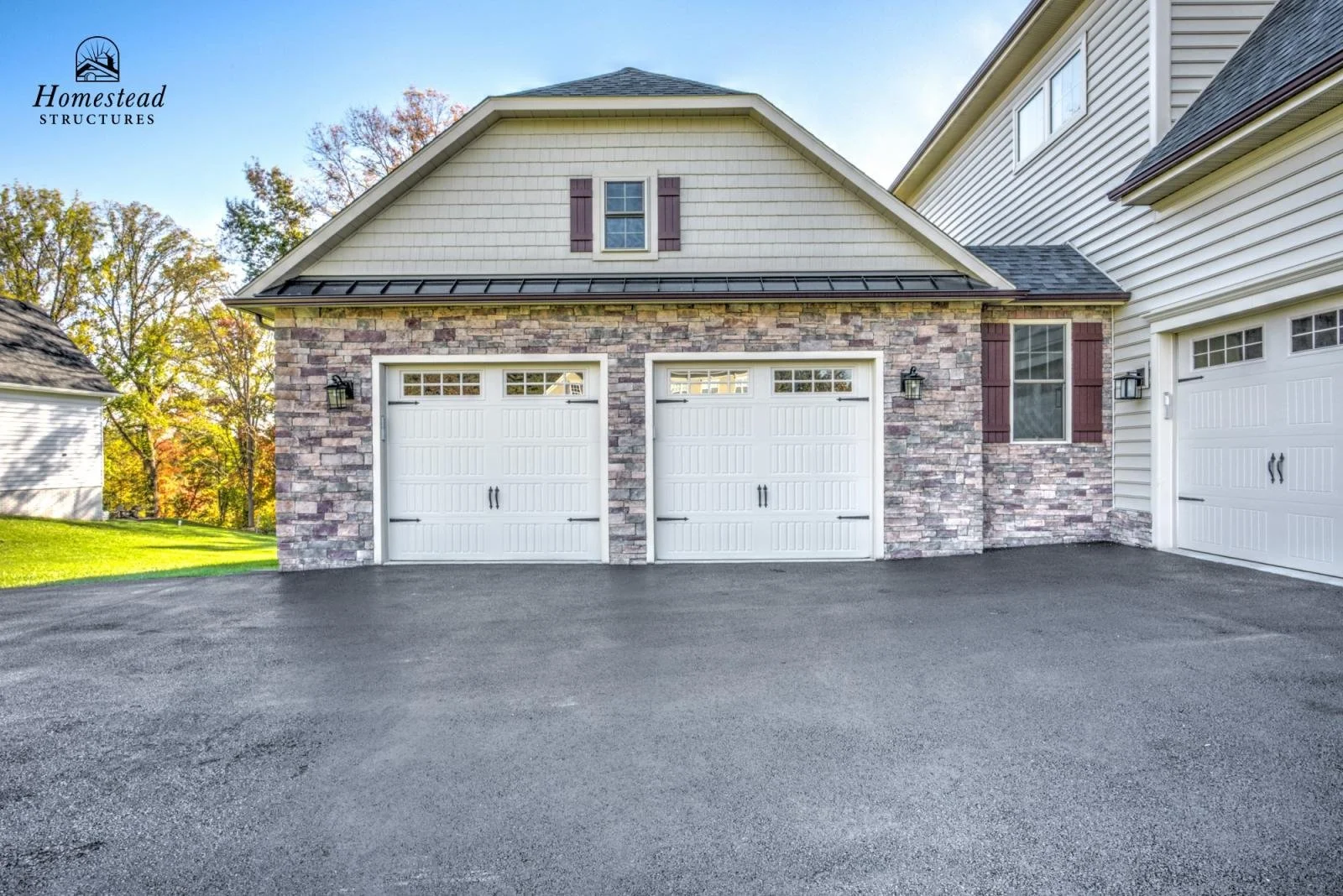 Newly built house with two white garage doors, brick and siding exterior, lamp fixtures, and windows, paved driveway, surrounded by trees with fall foliage.