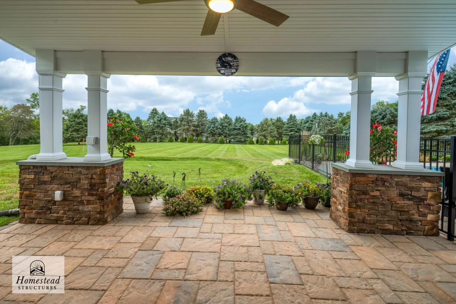 View of a backyard patio with a stone floor, white pillars with brick bases, potted plants, and a surrounding black metal fence, overlooking a well-maintained grassy area and trees under a partly cloudy sky.