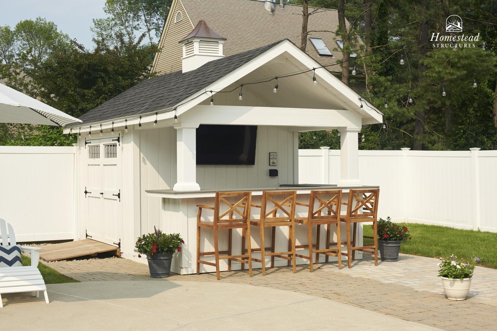 A white outdoor bar with a TV mounted on the wall, wooden barstools, and string lights hanging above, set in a backyard with a white fence and potted plants.