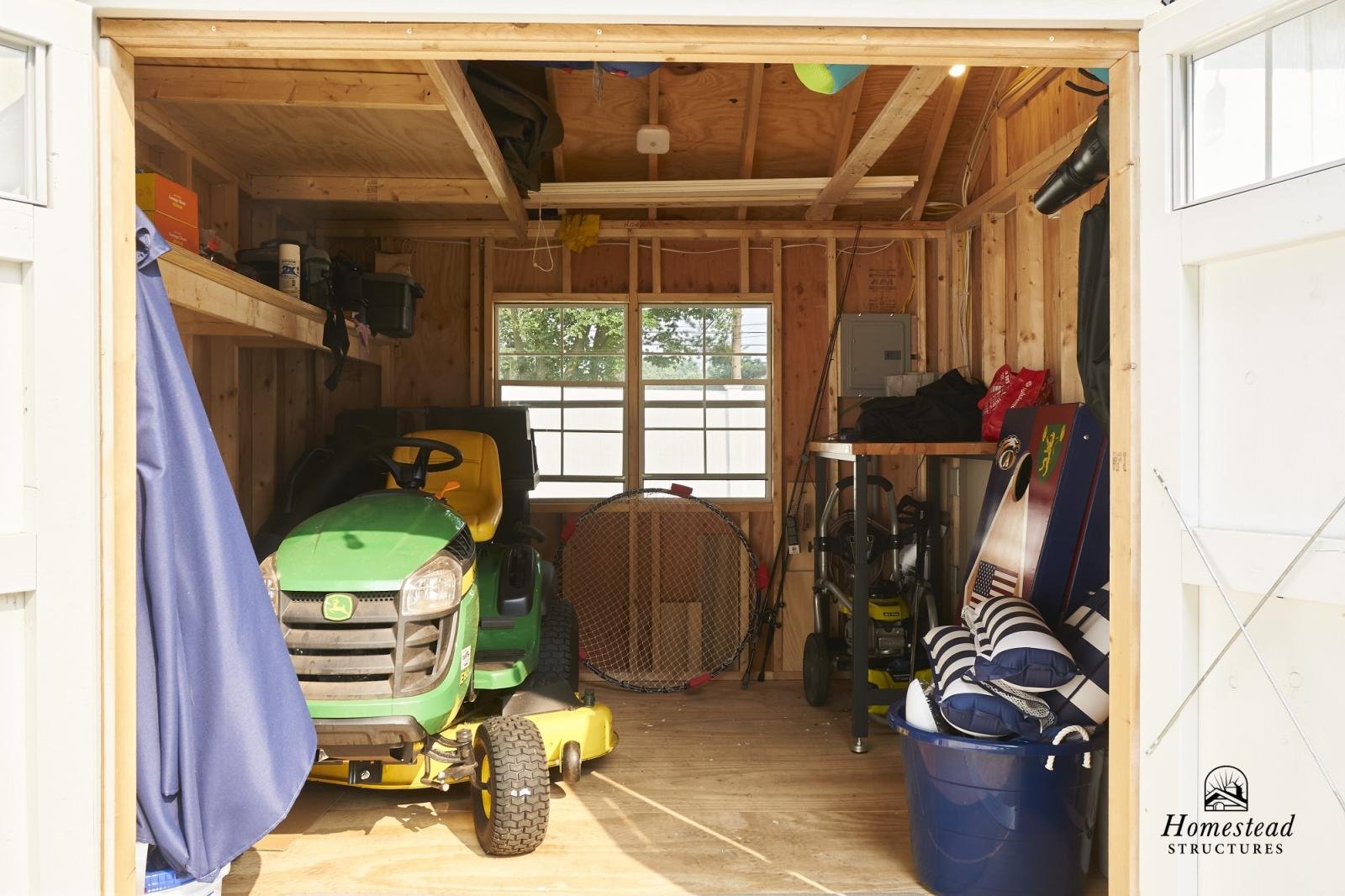 Inside a unfinished wooden garage, containing a green and yellow John Deere riding lawn mower, various outdoor tools, a blue storage bin, and shelving with boxes and supplies.