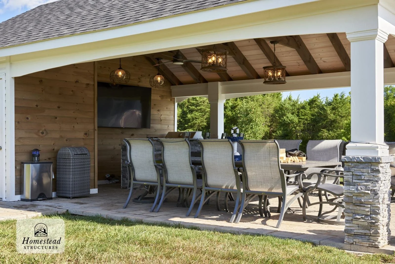 Covered outdoor patio with a large dining table and chairs, decorative hanging lights, a wall-mounted television, and a view of green trees in the background.