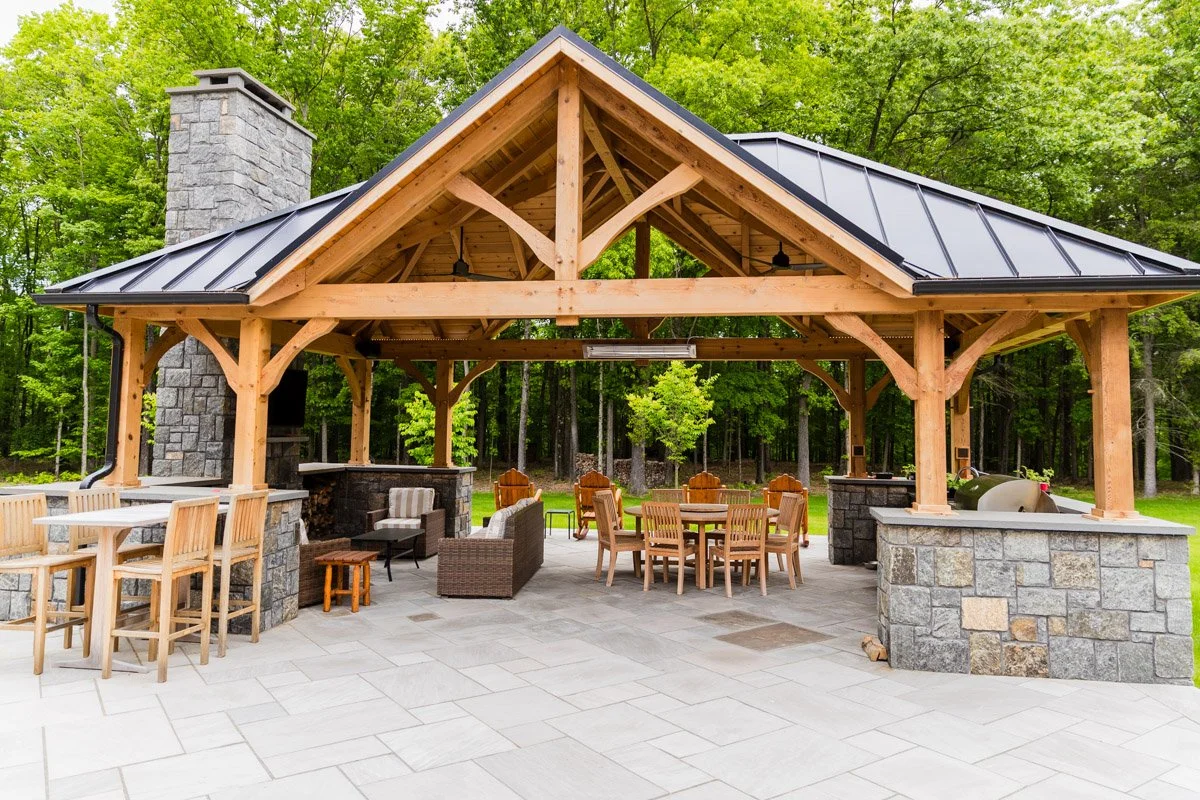 An outdoor pavilion with a wooden roof structure, stone pillars, and a chimney, surrounded by green trees. It has various outdoor furniture including bar stools, chairs, and tables, with a built-in grill and ceiling fans.