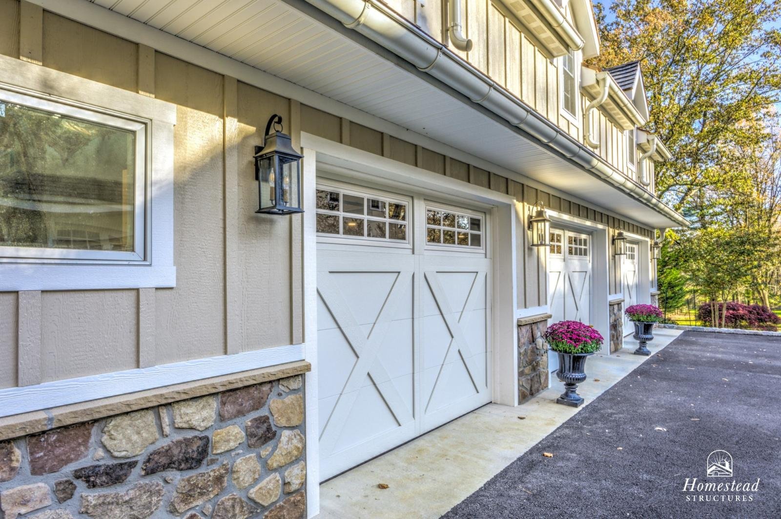 Exterior view of a house's garage with white doors, stone and wood siding, black lantern-style light fixtures, and pink flower pots along the driveway, surrounded by trees with autumn foliage.