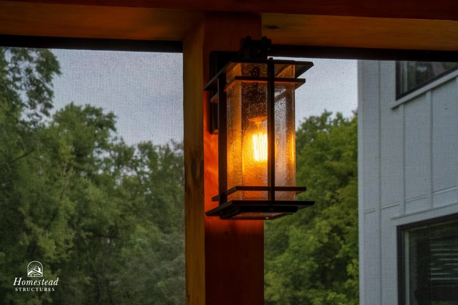 Outdoor porch lantern with metal frame and glass panels, illuminated at dusk with trees and house in the background.