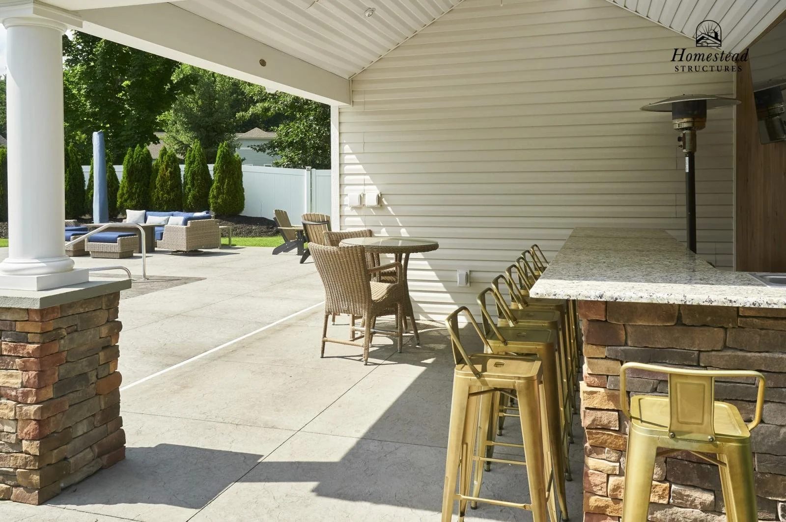 Outdoor patio area with bar seating, granite countertop, and wicker chairs under a roof, with a seating area featuring cushioned chairs and lounge chairs with umbrellas in the background.