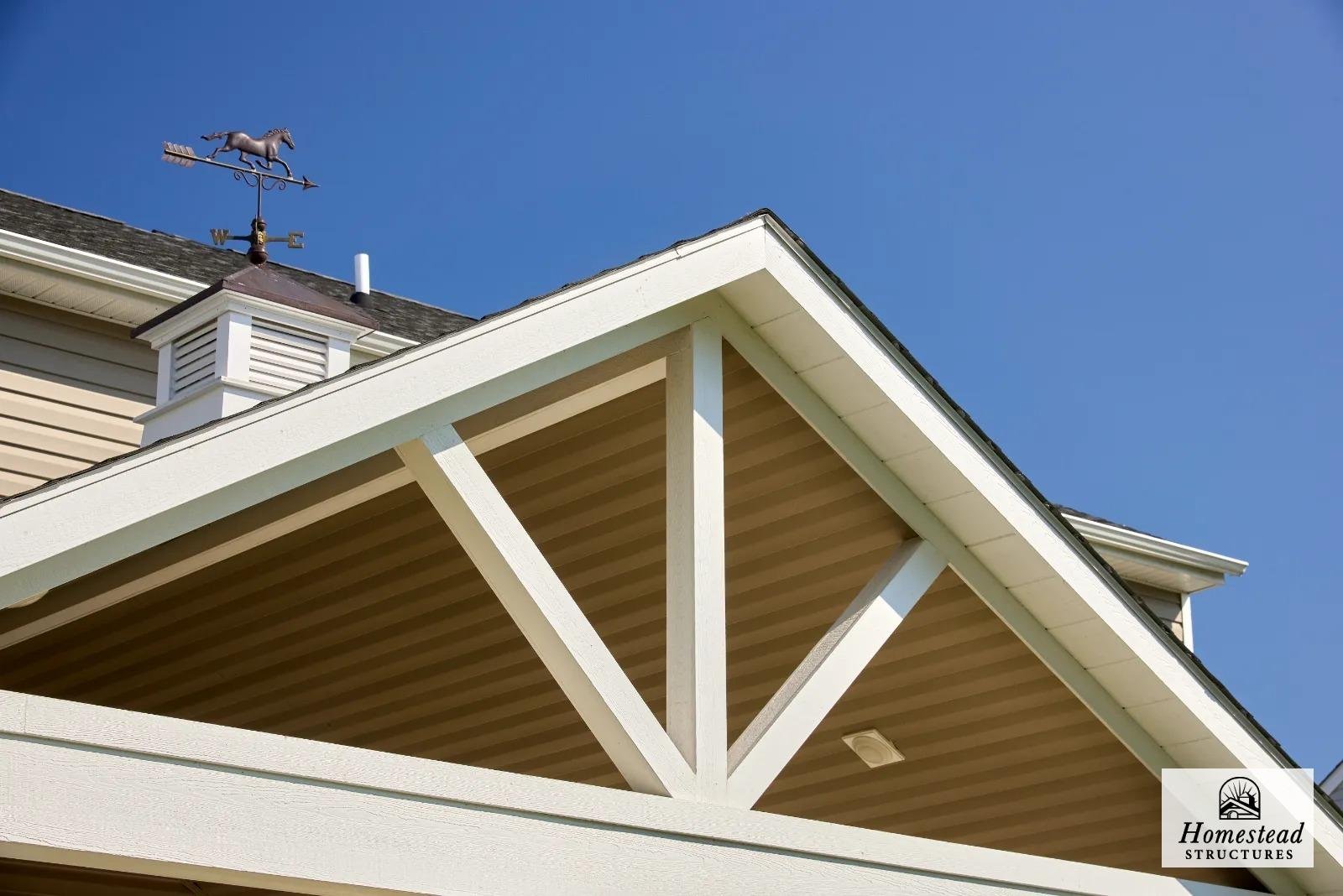 View of the underside of a house's porch roof with white painted beams, beige siding, and a blue sky in the background.