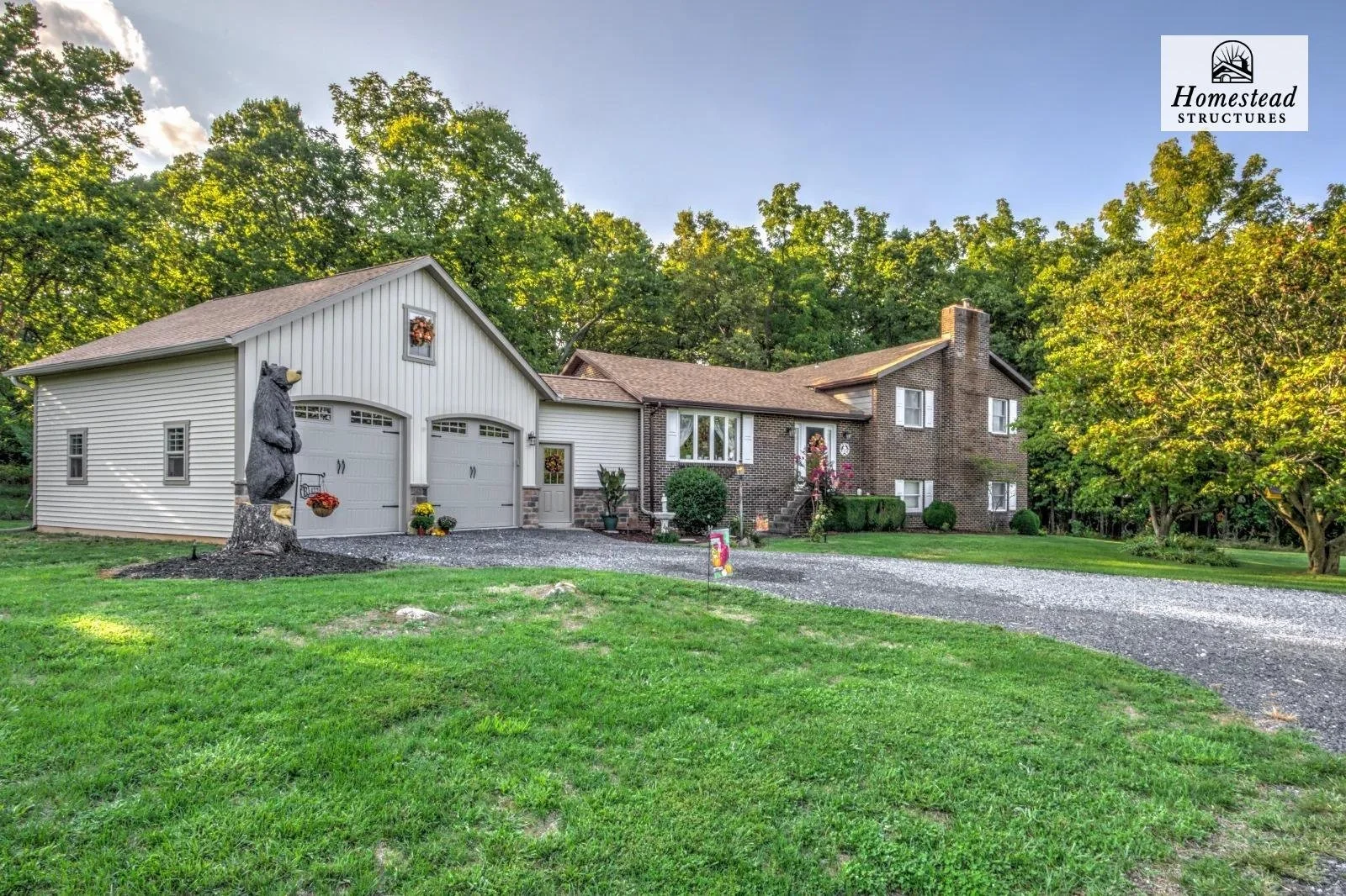 A house with a garage and a large lawn, decorated with autumn and Halloween decorations, surrounded by trees, with a gravel driveway leading up to the house.