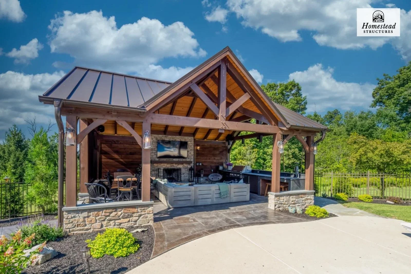 Outdoor pavilion with wooden beams and a metal roof, featuring a fireplace, seating area, and outdoor kitchen, surrounded by a landscaped yard and trees, under a partly cloudy sky.