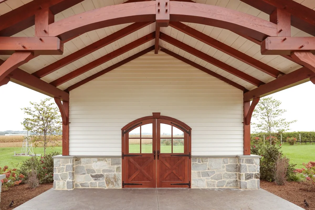 View of a barn with wooden doors, stone foundation, and white siding, set in a rural landscape with green grass and trees.