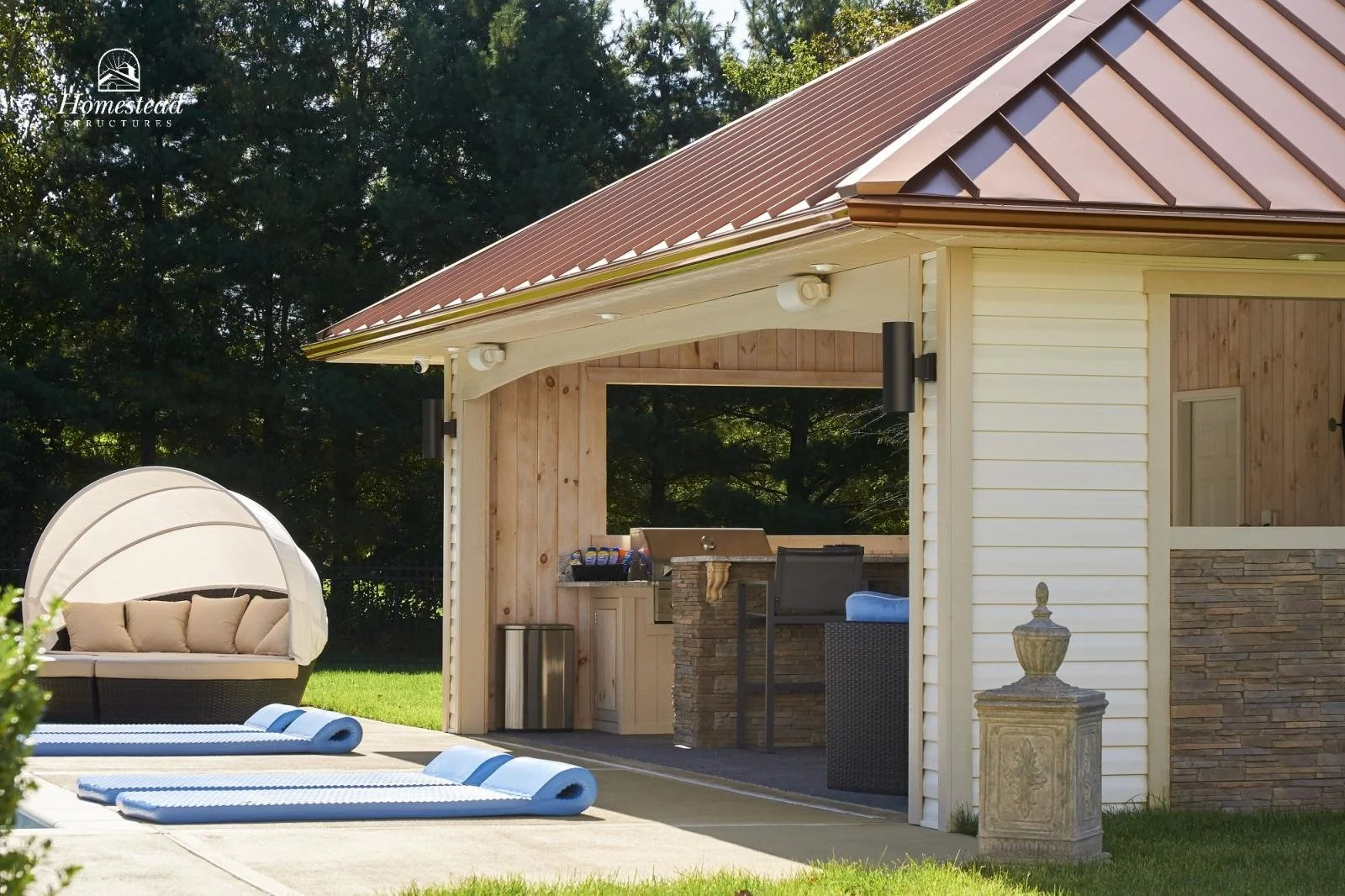 Outdoor backyard patio with a covered area featuring a barbecue grill, high-top counter with barstool, and an upholstered daybed with cushions, on a sunny day.