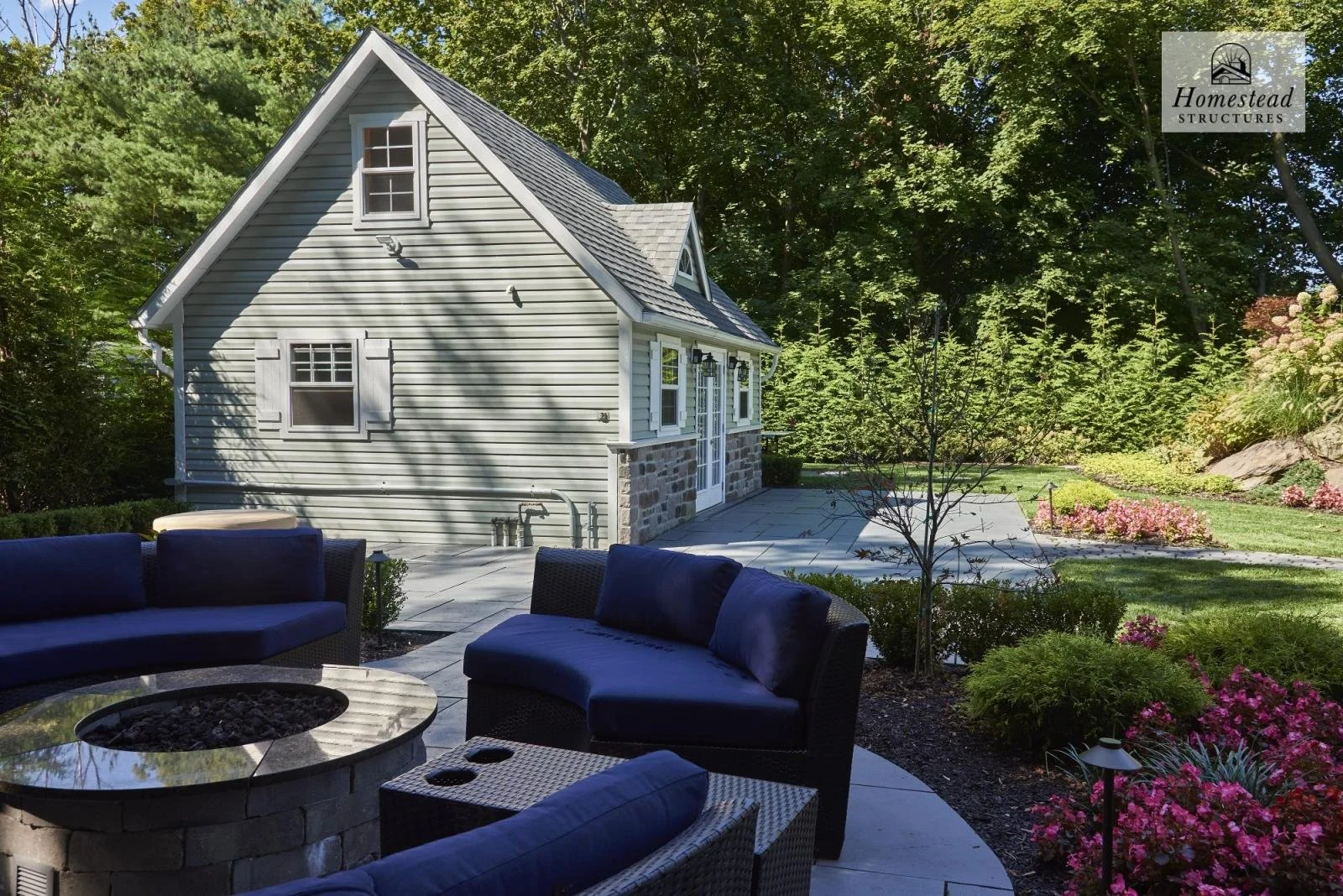 A backyard scene featuring a small house with gray vinyl siding, white window shutters, and a stone foundation. In the foreground, there is outdoor furniture including a fire pit and cushioned chairs, with a walking path, landscaped flowers, shrubs, 