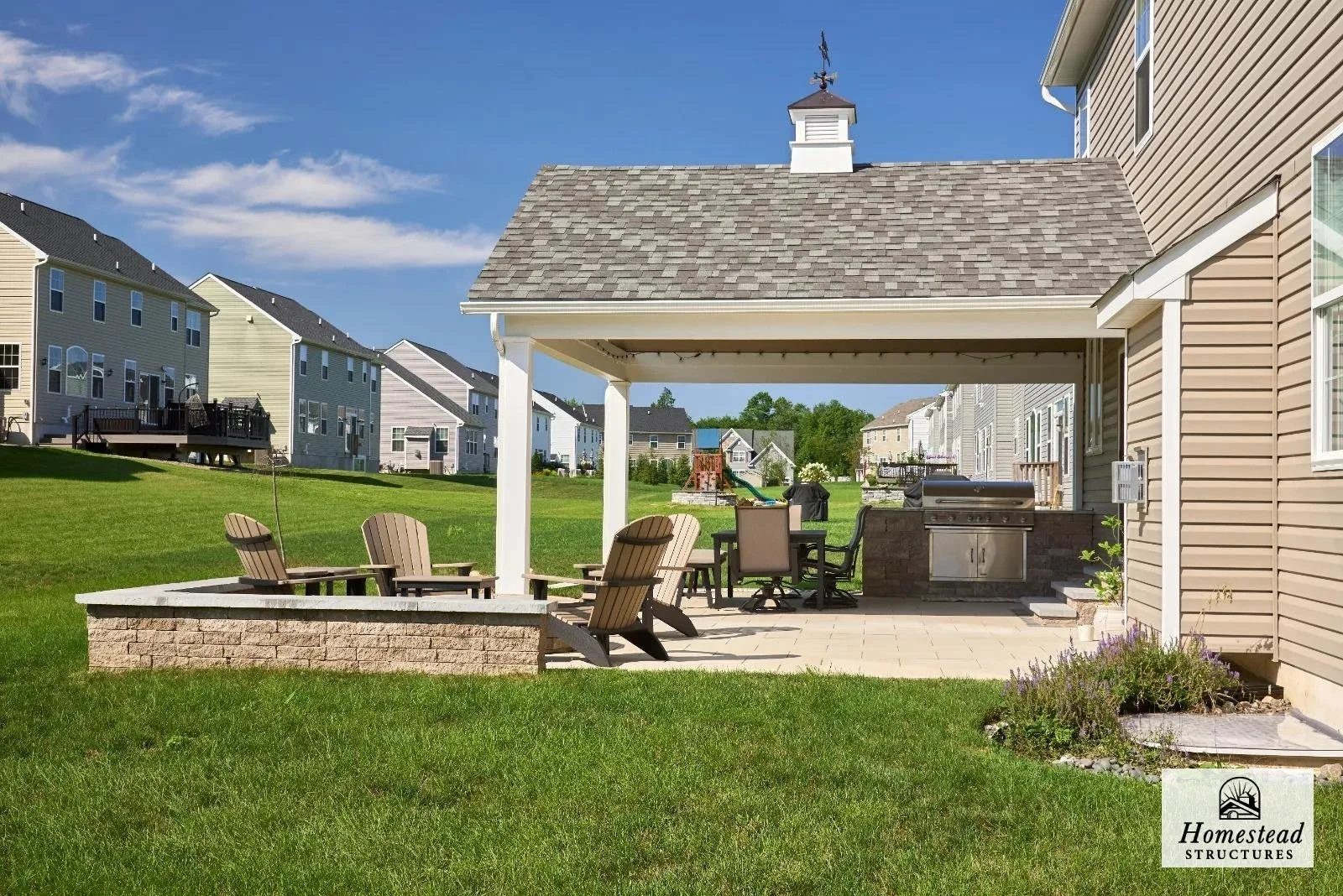 Backyard patio with Adirondack chairs, a table, and a barbecue grill under a covered extension with a small cupola on roof, surrounded by a grassy yard and neighboring houses.