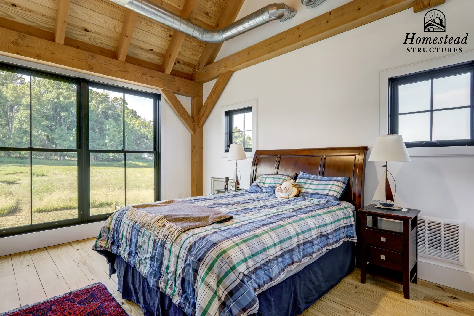 Bedroom with a bed, plaid bedding, wooden headboard, two lamps, a small decorative sailboat, a window, small black window, and large window showing greenery outside.