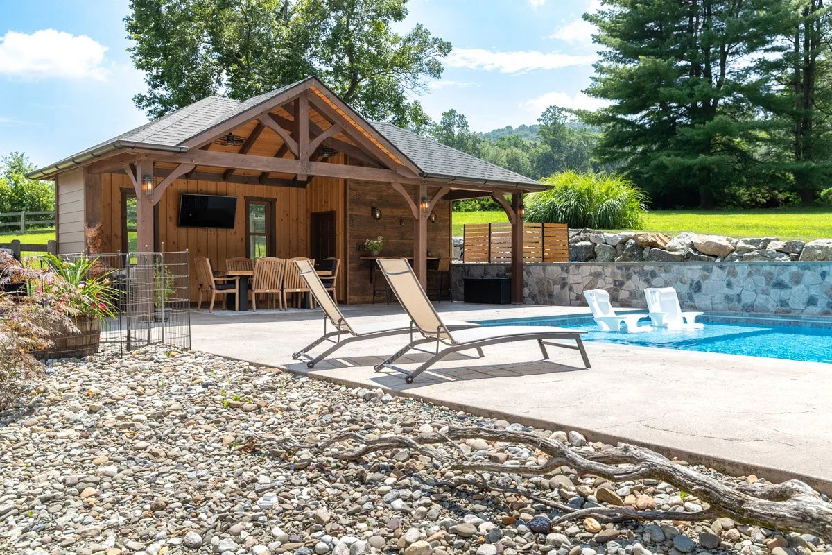 Backyard with a swimming pool, two lounge chairs, a wooden cabana, a seating area, a TV, and lush green trees in the background.