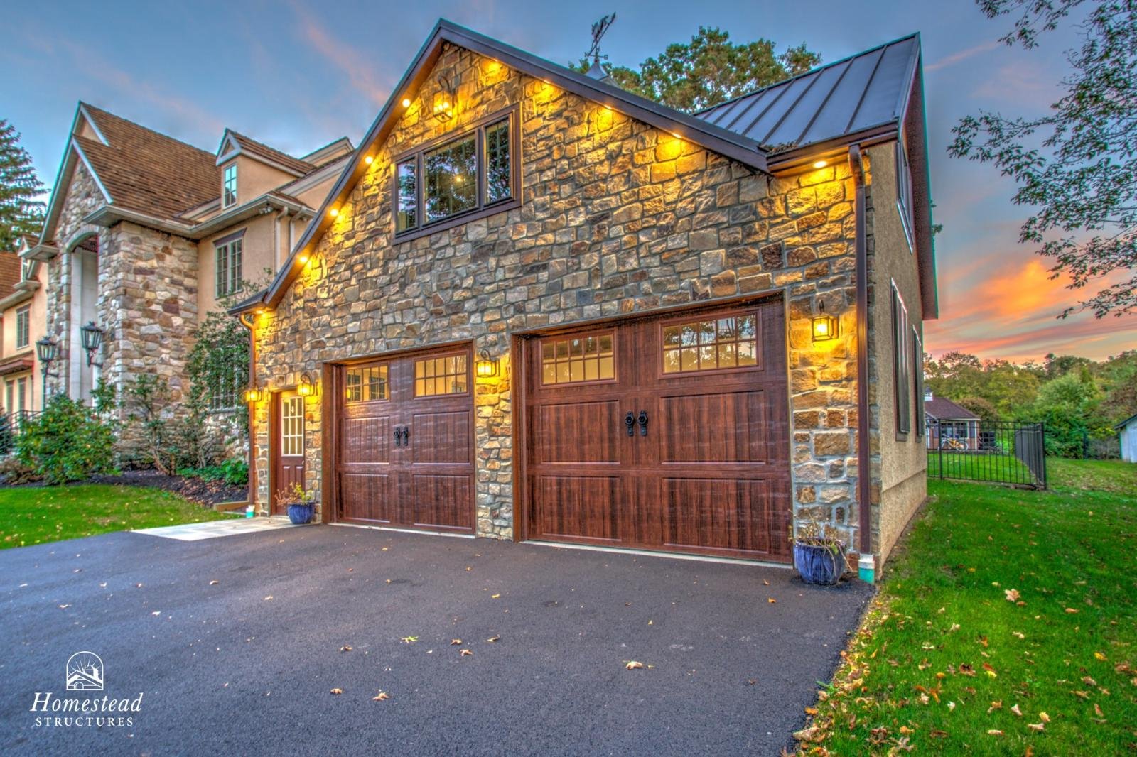 A modern house with stone and stucco exterior, two wooden garage doors, porch lights, and a small side door, surrounded by greenery and a sunset sky.