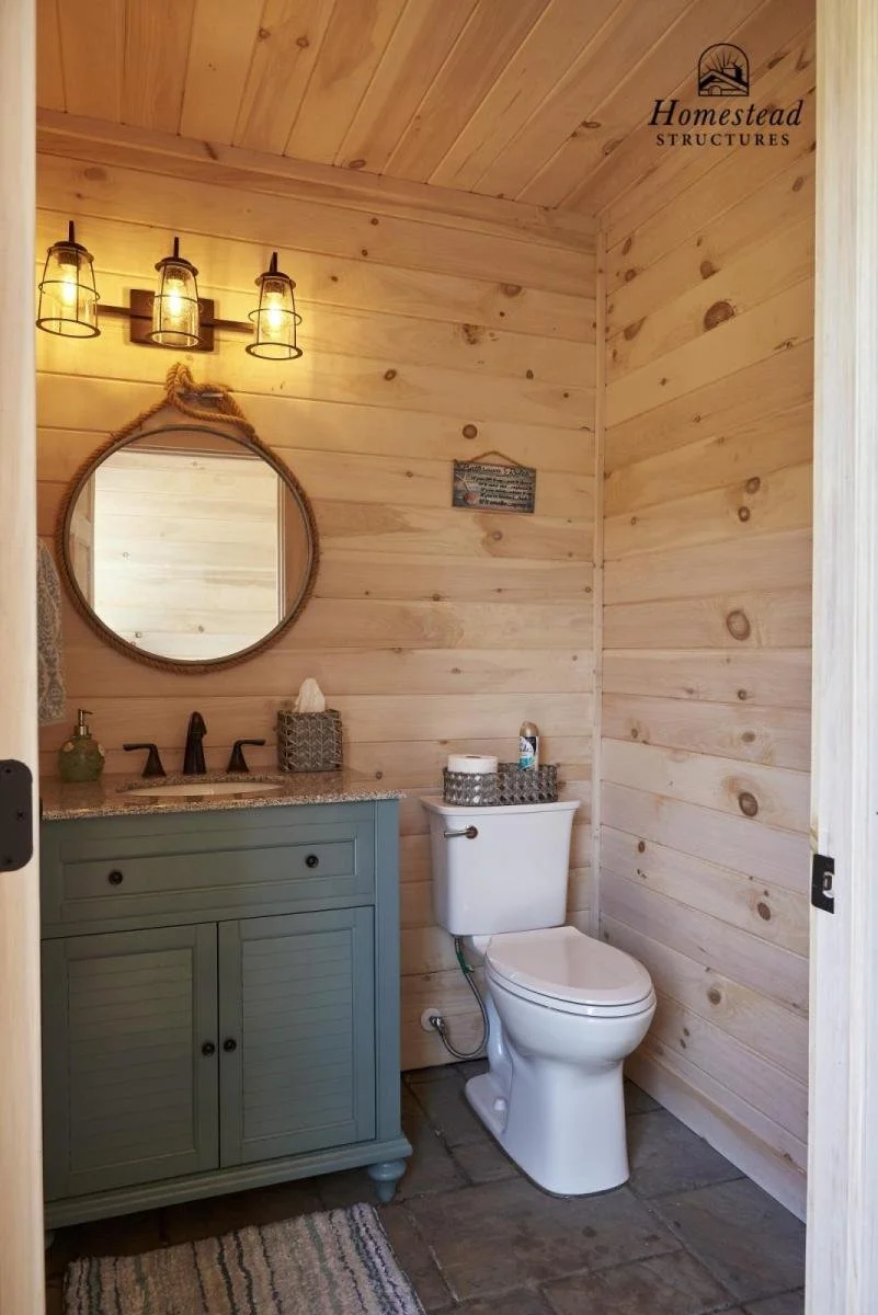 A rustic bathroom with light wood paneled walls and ceiling, a round mirror hanging above a green vanity with a granite countertop, a black faucet, and a soap dispenser. A three-light industrial-style wall sconce is mounted above the mirror. There is