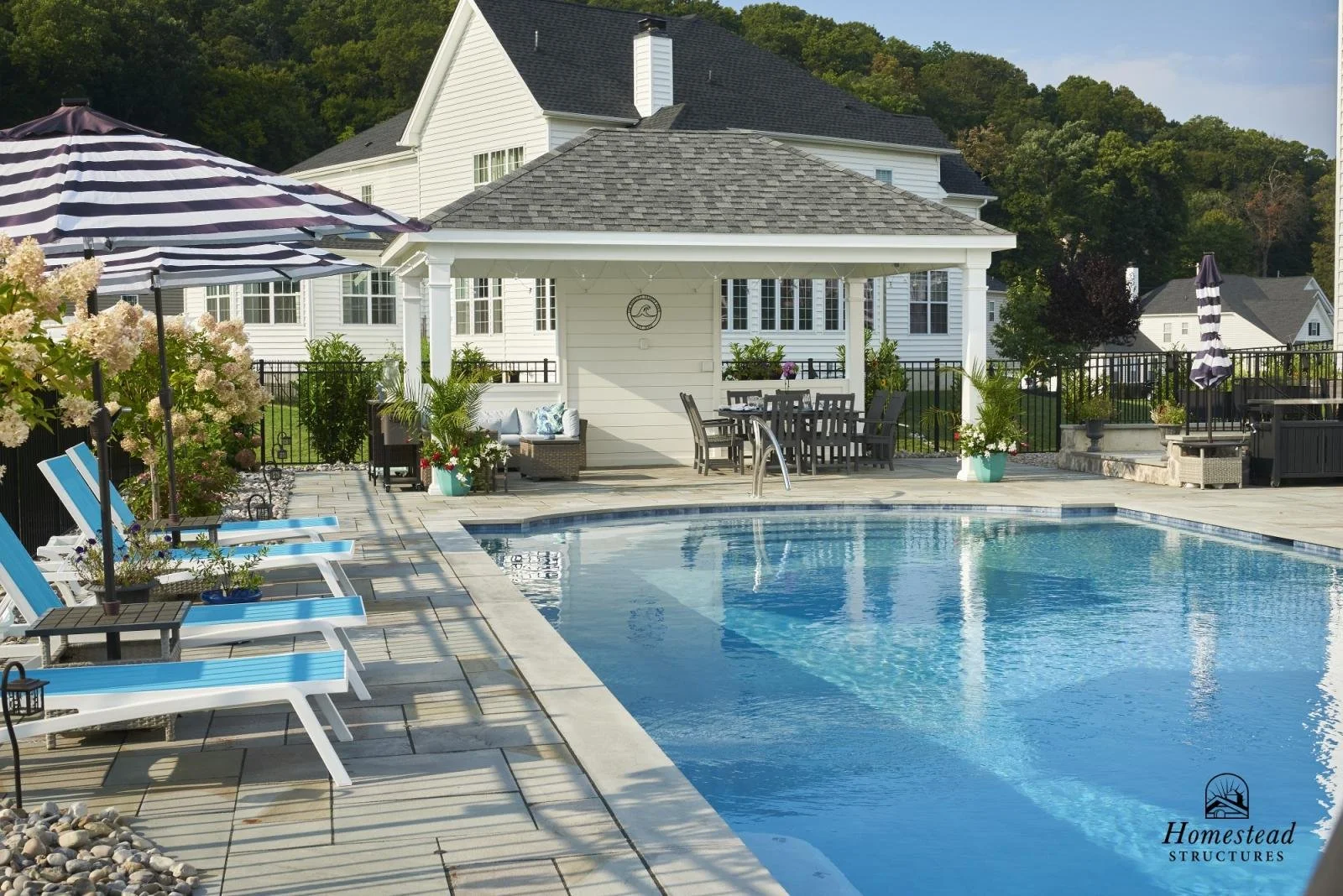 A backyard swimming pool with lounge chairs, umbrellas, and a pool house in the background, surrounded by greenery and neighboring houses.