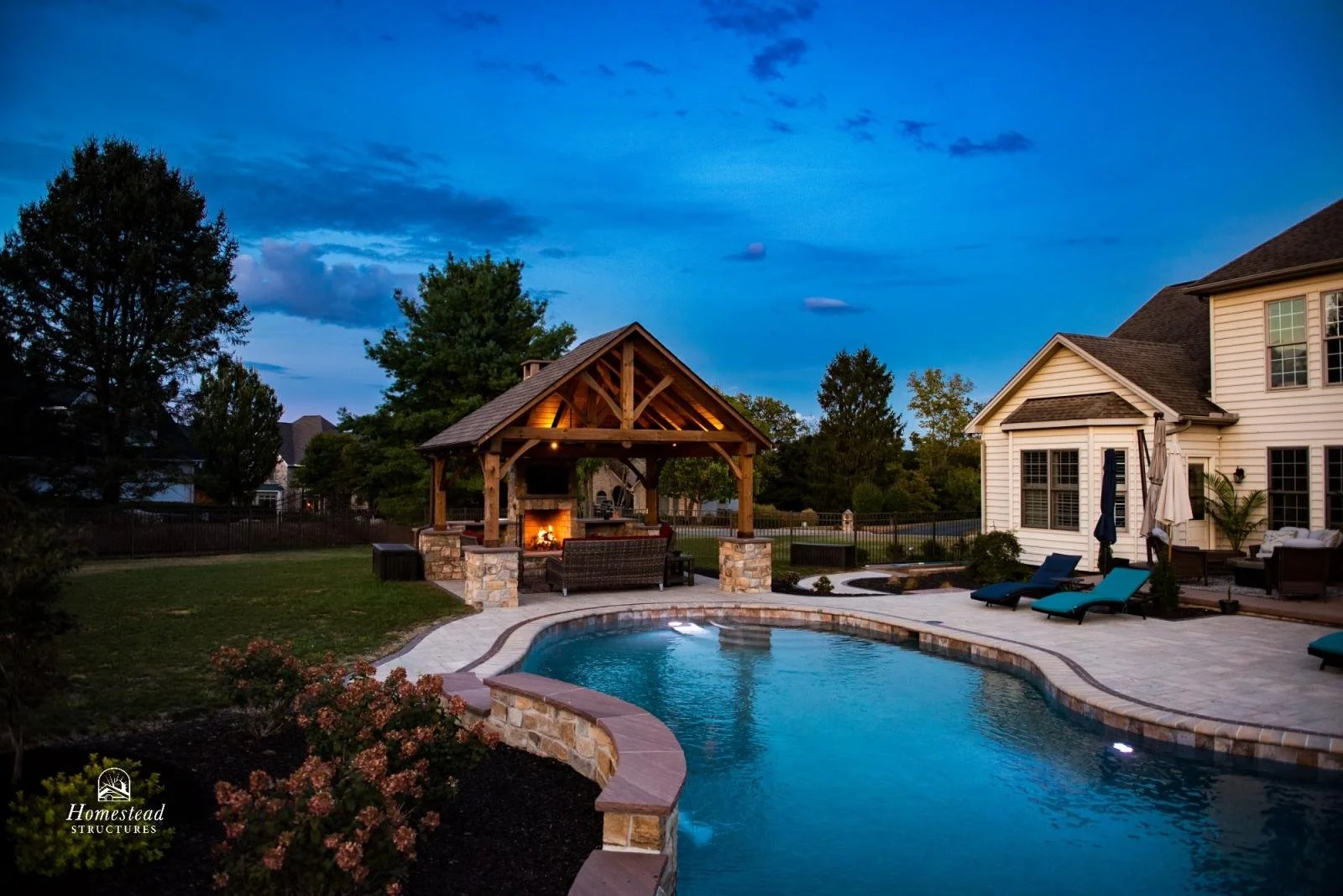 Beautiful backyard with a swimming pool, a wooden outdoor pavilion with a fireplace, outdoor seating, lounge chairs, and a covered patio area with a house visible in the background during dusk.