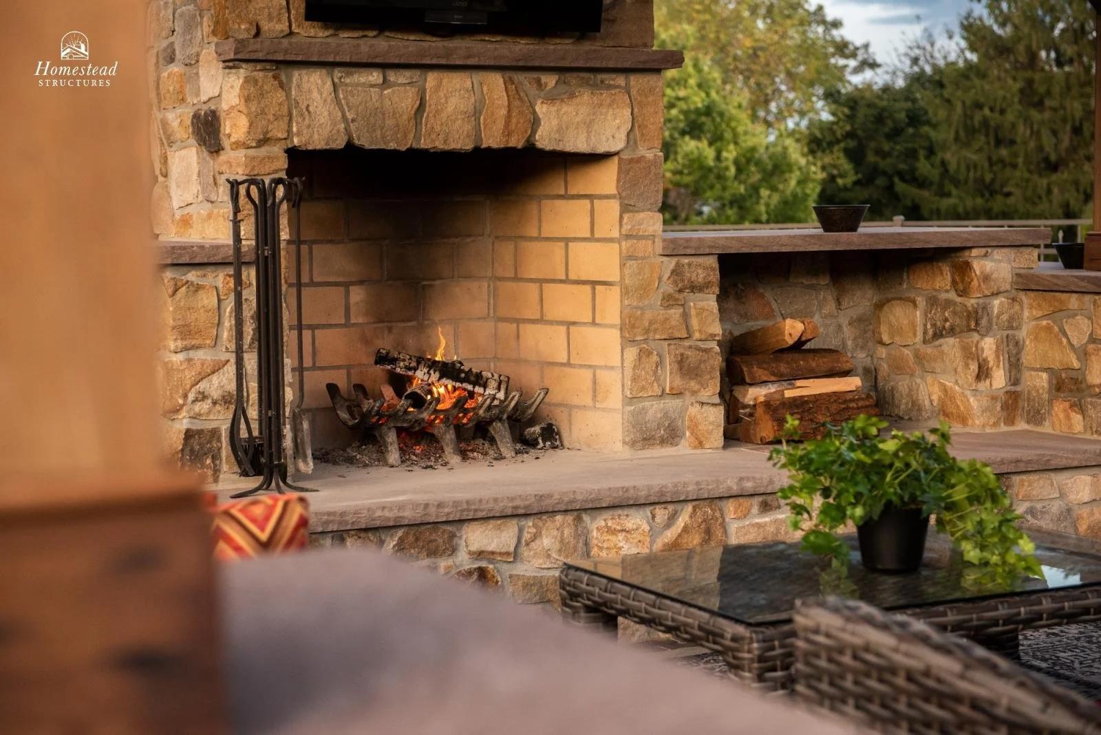 Outdoor stone fireplace with burning logs and a fire grate, surrounded by a stone wall, with chopped firewood stacked nearby, and a wicker table with a green potted plant, set in a patio area with trees in the background.