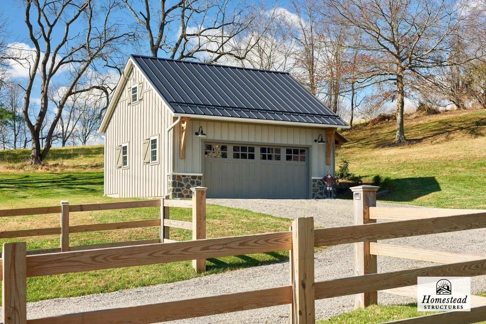 A small, modern garage with beige vertical siding, a black metal roof, and a stone foundation, situated on a grassy hill with trees in the background. There is a gravel driveway leading to the garage, and a wooden fence in the foreground. The sky is 