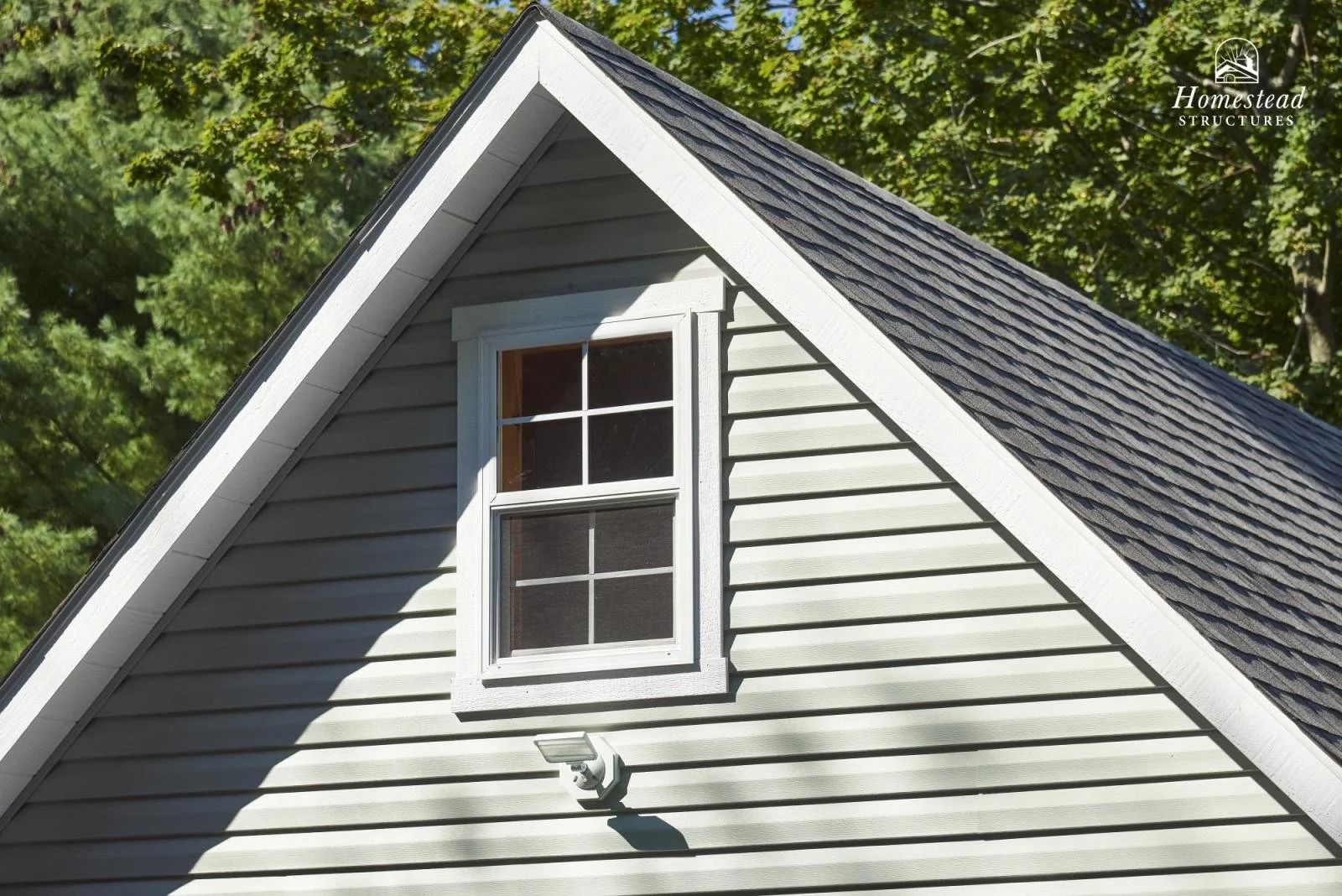 Close-up of a house's gable roof with gray shingles, white siding, a double-hung window, and a security light post against a background of green trees.