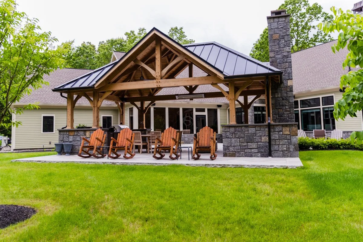 Backyard patio with wooden pergola, Adirondack chairs, and patio furniture, surrounded by green grass and trees.