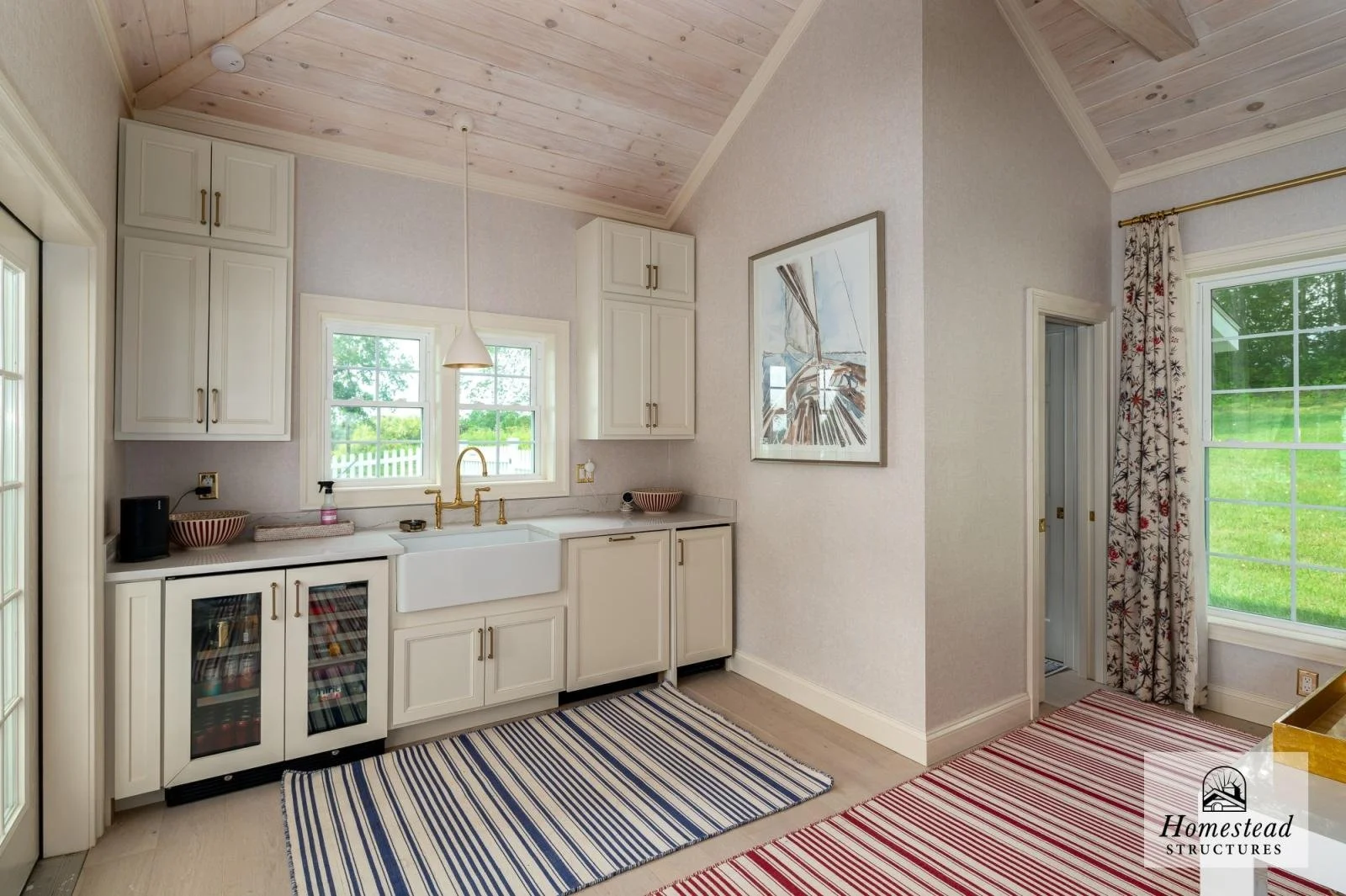 Clean, bright kitchen with white cabinets, a farmhouse sink, a window above the sink, and a wood-paneled vaulted ceiling. There are two rugs on the light-colored floor, one striped in blue and white, the other striped in red and white. To the right, 