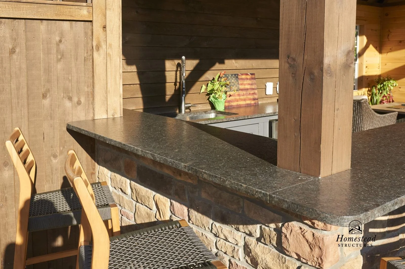 Outdoor kitchen with a stone and wooden counter, a sink, and chairs.