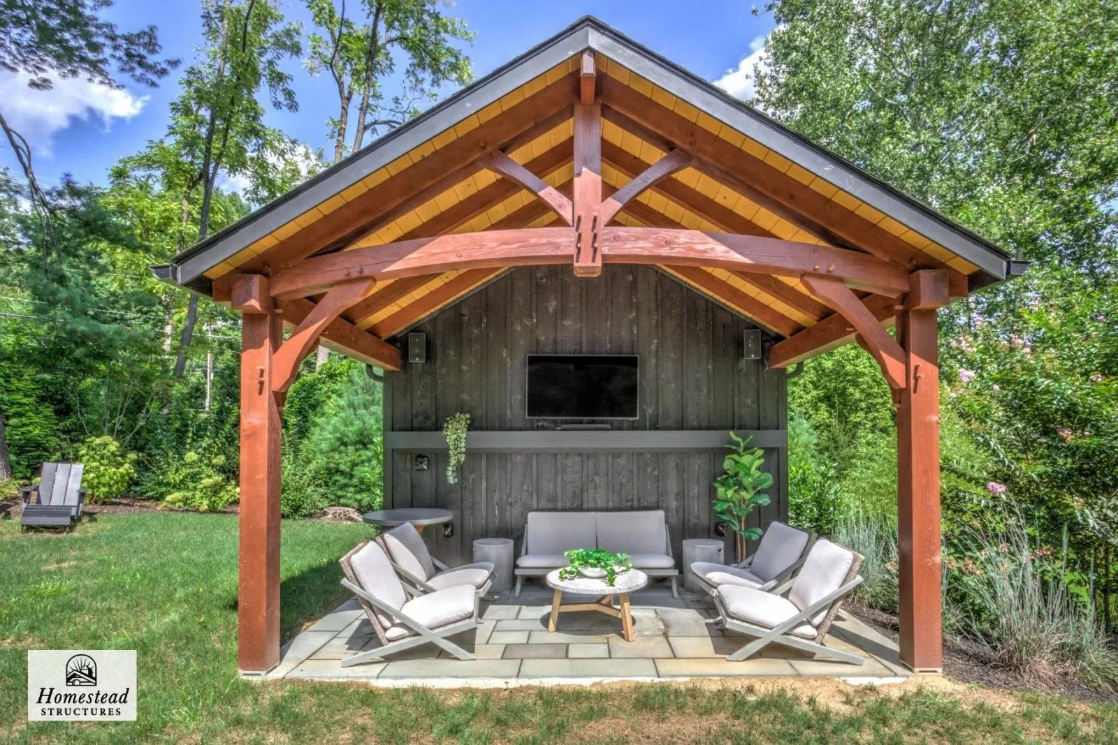 Outdoor wooden pavilion with a pitched roof, black wall, mounted TV, and six beige cushioned chairs arranged around a small coffee table in a lush green garden.