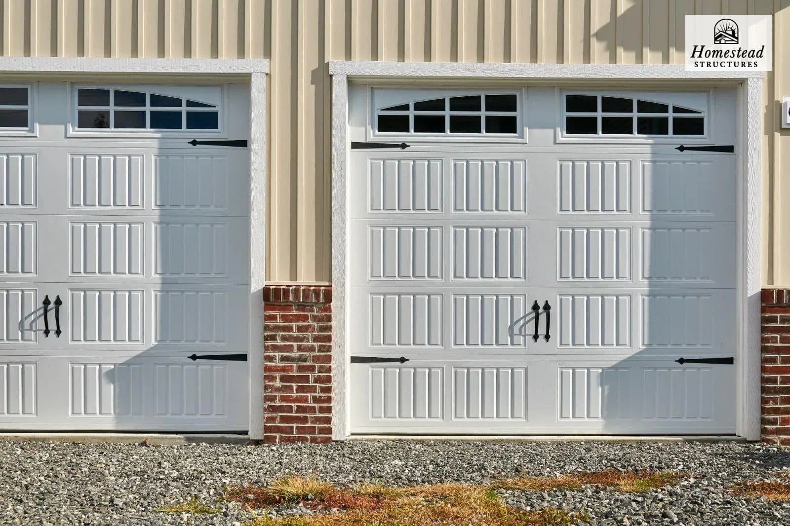 Two white garage doors with black hardware and small windows at the top, set against a beige metal building with brick accents. "Homestead Structures" logo is in the top right corner.