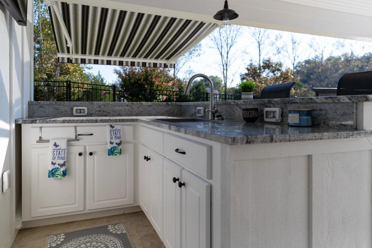 Outdoor kitchen with white cabinets, granite countertop, sink, and a retractable striped awning. Decorated with a potted plant and small containers, with trees and fence in the background.