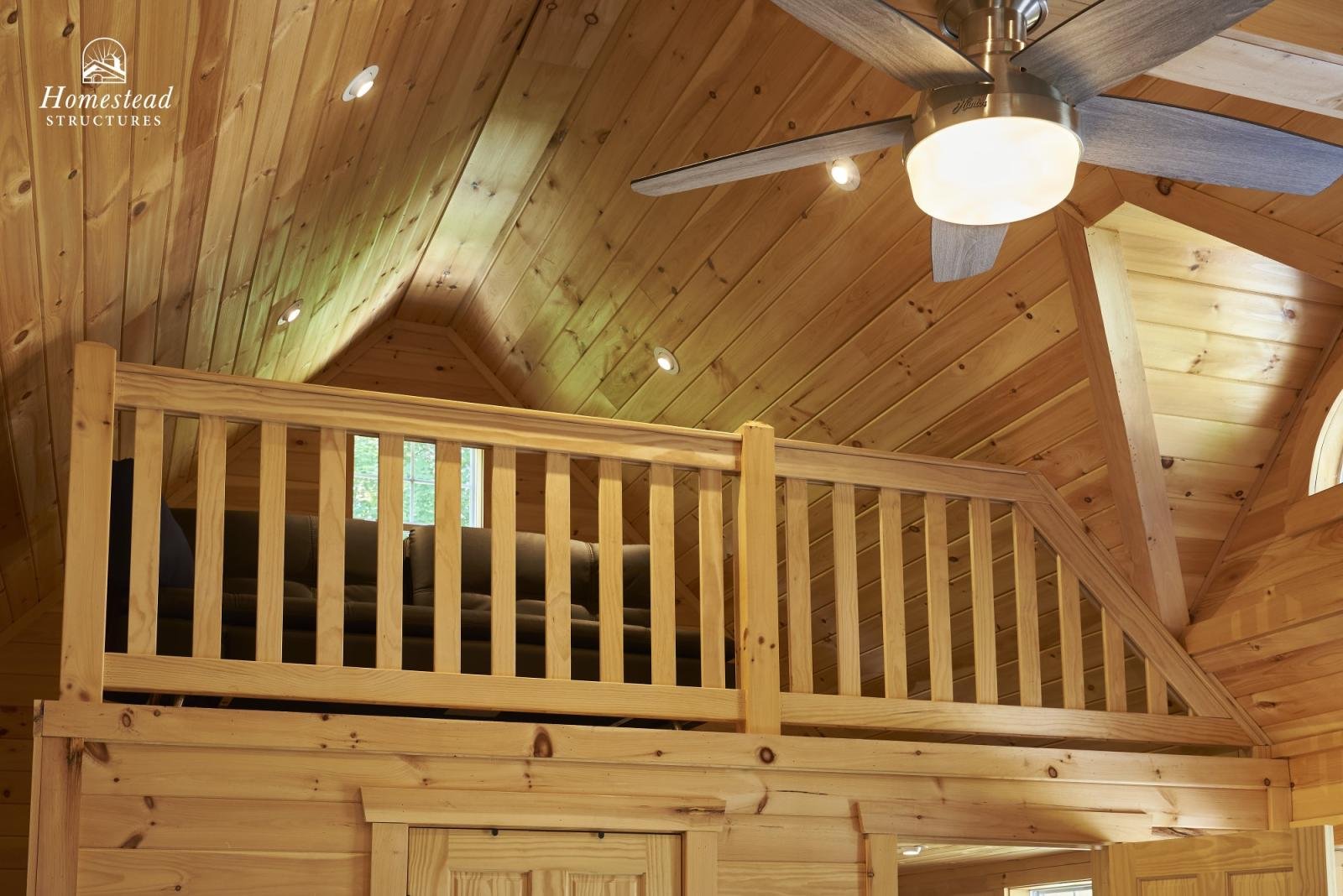 Interior view of a wooden cabin with a lofted area, wooden paneling on the walls and ceiling, a ceiling fan with lights, and small windows.