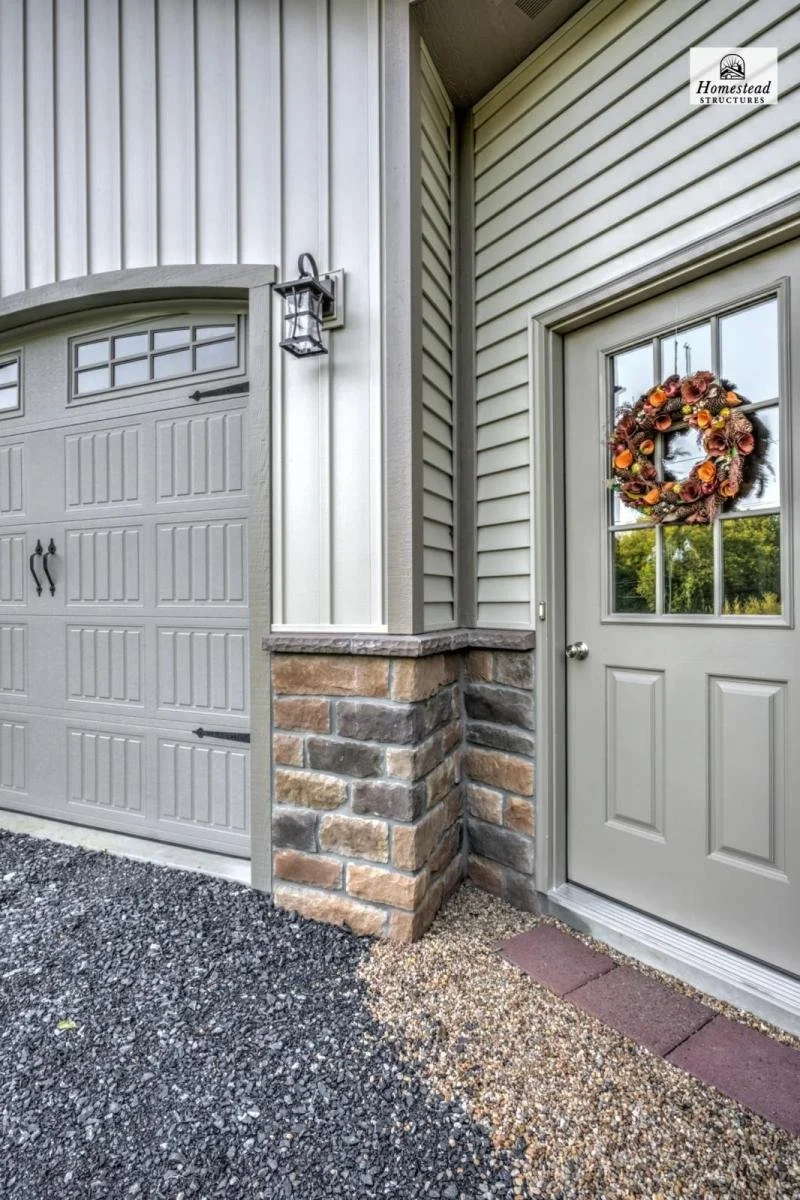 Exterior corner of a house showing a garage door, a wooden door with a wreath, a lantern, siding, and brick foundation, with gravel and stone ground.