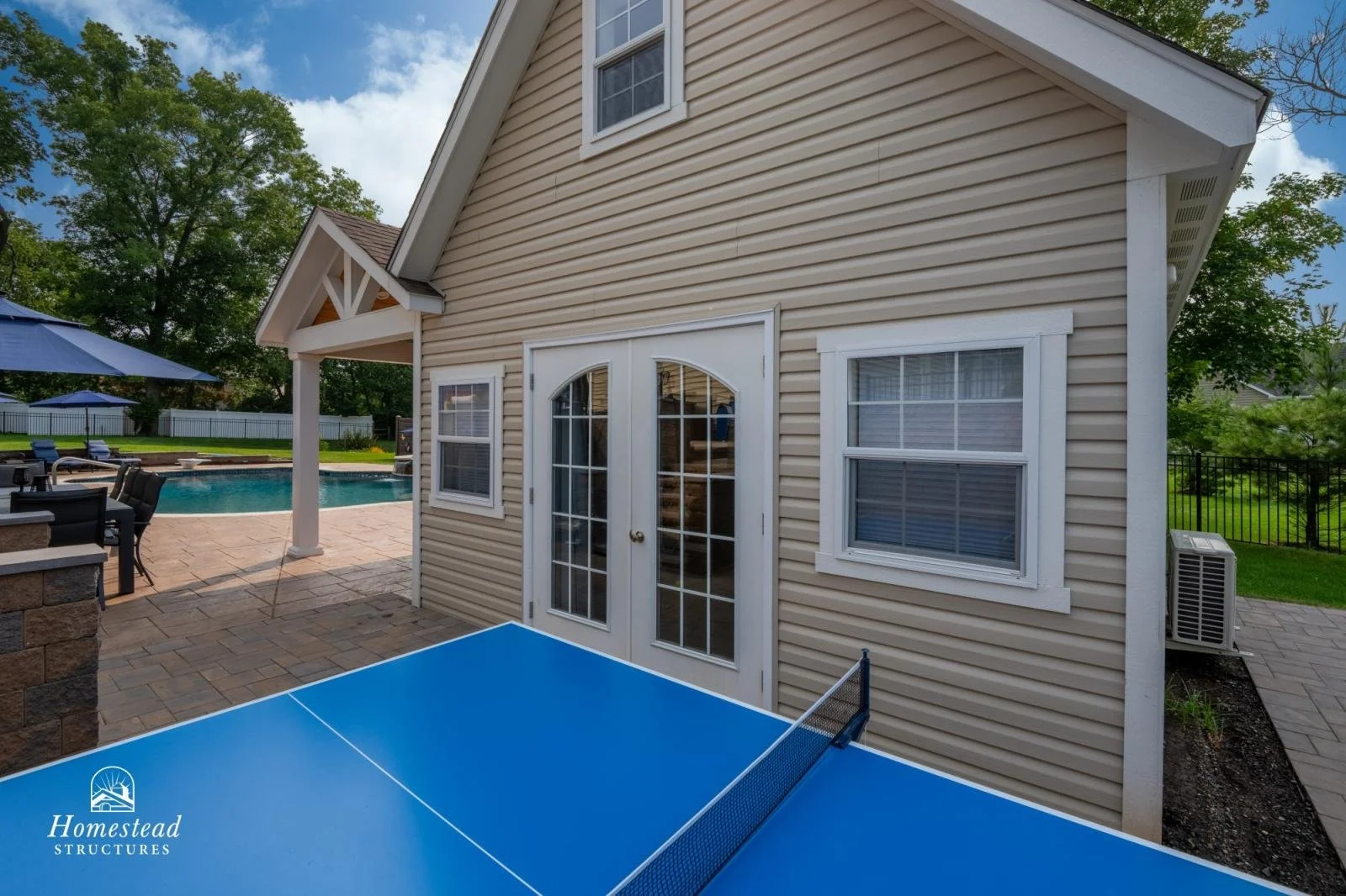 Backyard with pool, outdoor dining area, and pool house with beige siding and white French doors.