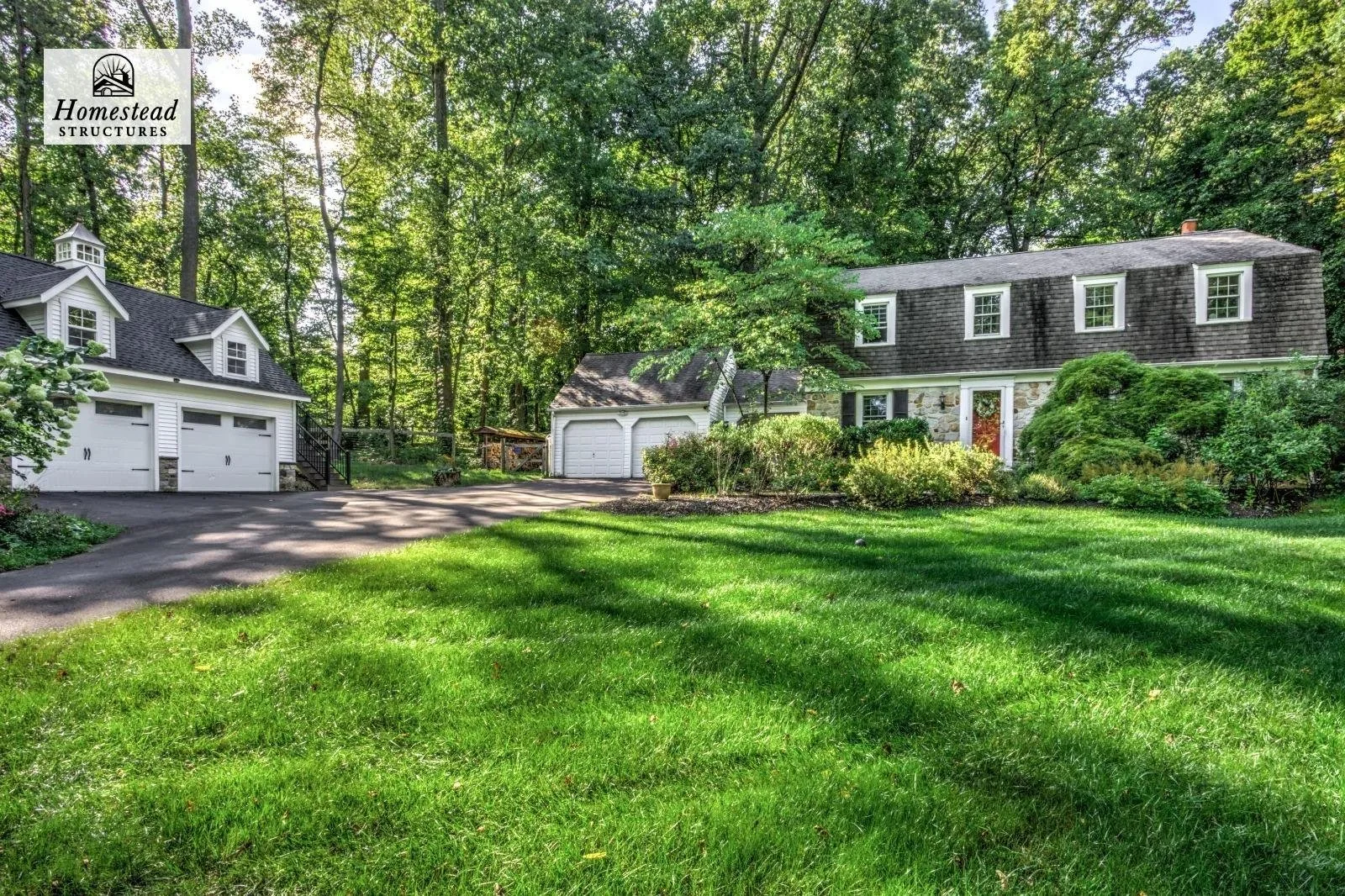 Front yard of a house with a green lawn, trees, shrubs, and a paved driveway leading to a separate garage, with a wooded area in the background.