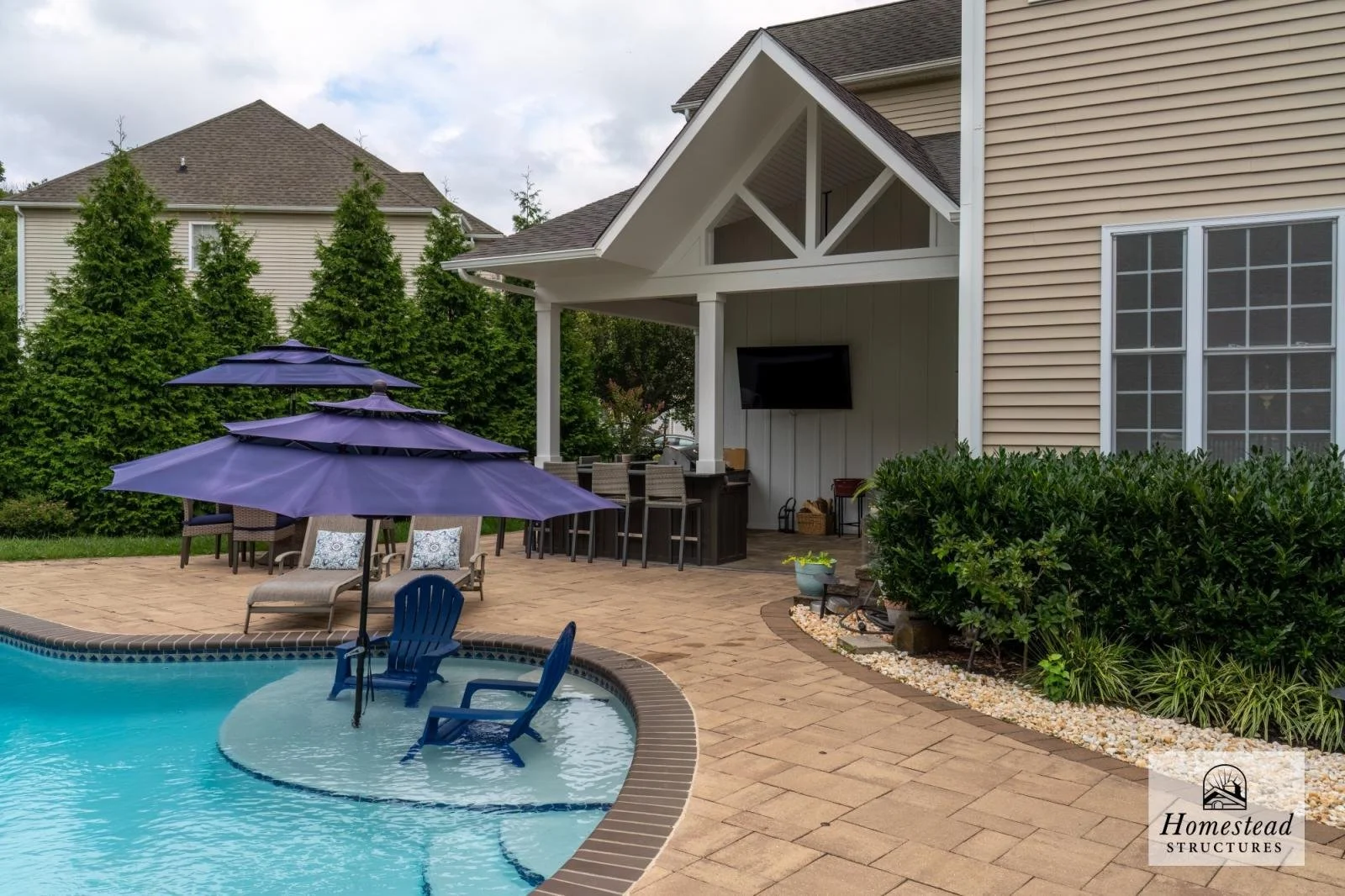 Backyard with swimming pool, four purple umbrellas, lounge chairs, a bar area with stools, a mounted TV, and green shrubbery.
