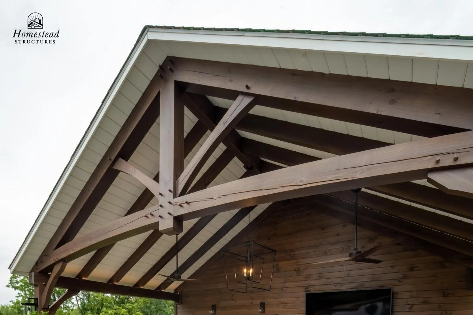 Close-up of a wooden house roof with exposed beams, white siding, a hanging lantern, ceiling fans, and a flat-screen TV on the wooden wall, with greenery visible in the background.