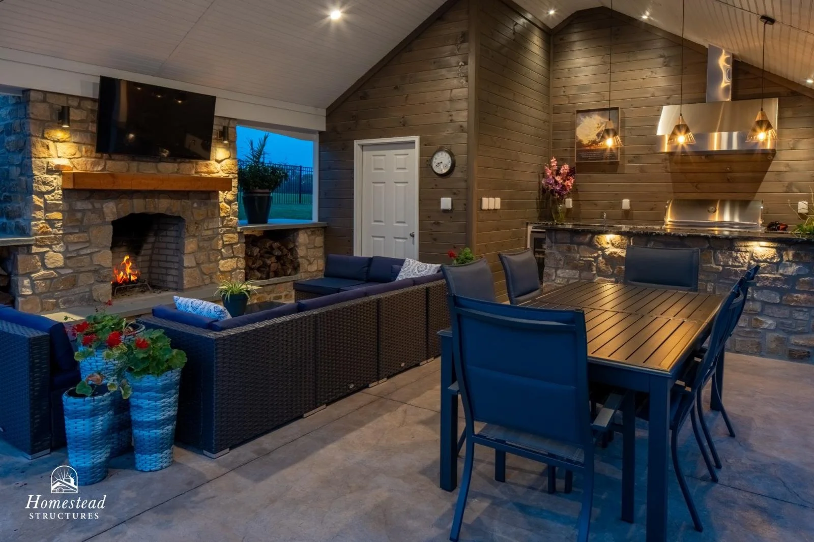 An outdoor living space with a stone fireplace, a flat-screen TV, and a wood-paneled ceiling. There are cozy couches with cushions, a dining table with chairs, potted plants, and an outdoor kitchen area with a stone counter and stainless steel applia